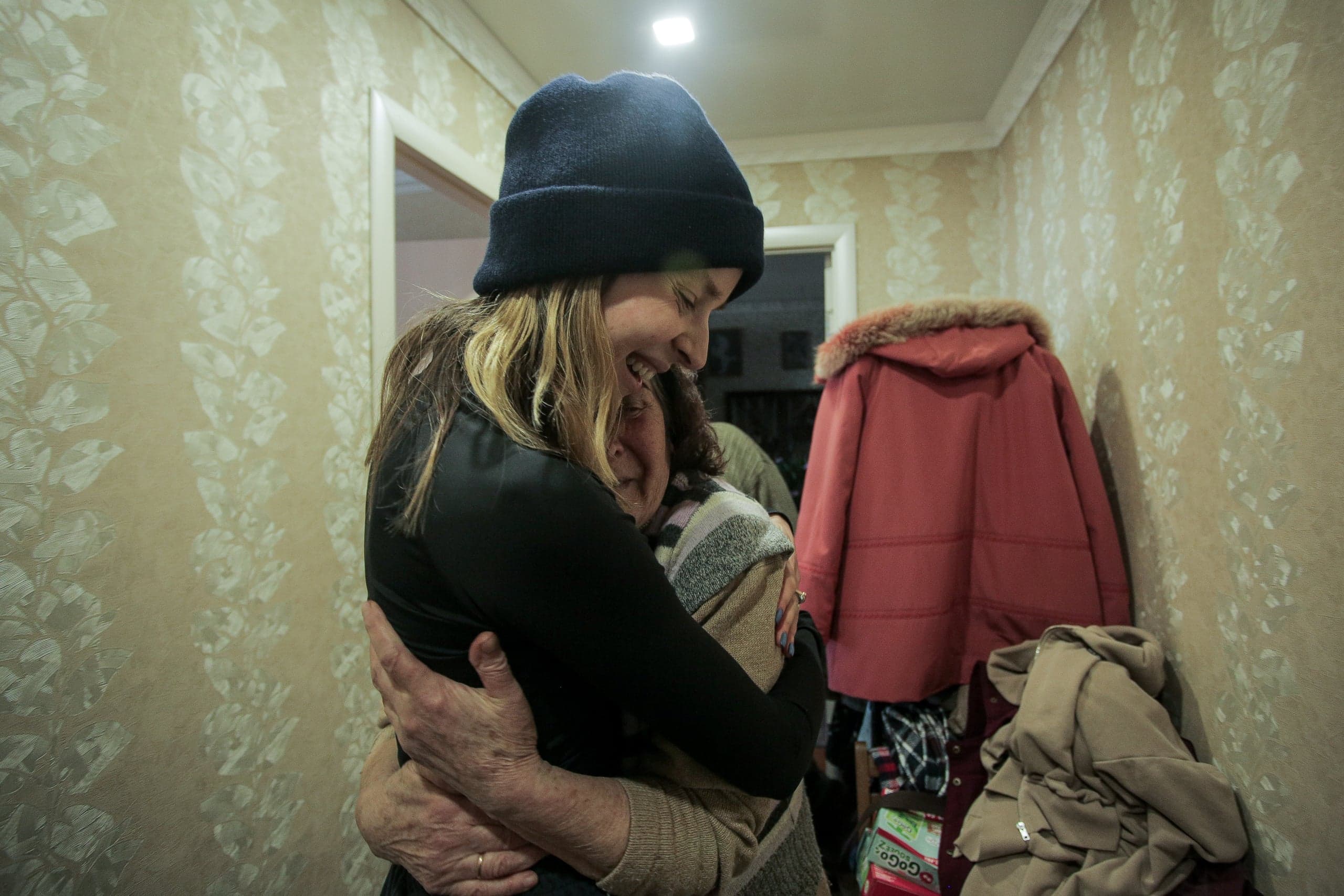 Крепкие объятия двух женщин в прихожей с обоями. Женщина в черной шапке улыбается.