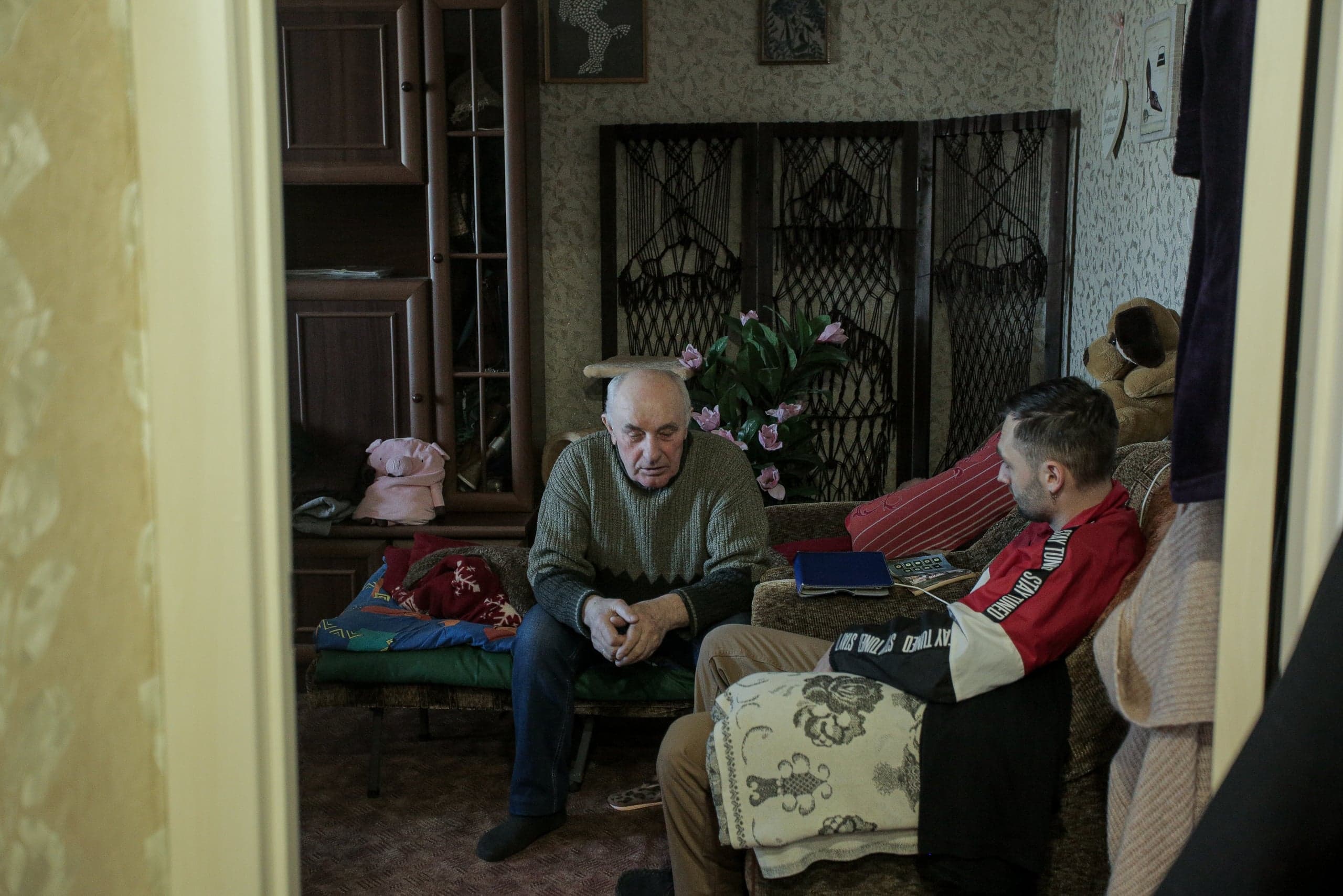 Пожилой и молодой мужчины сидят в тускло освещенной комнате среди старой мебели и домашней утвари.