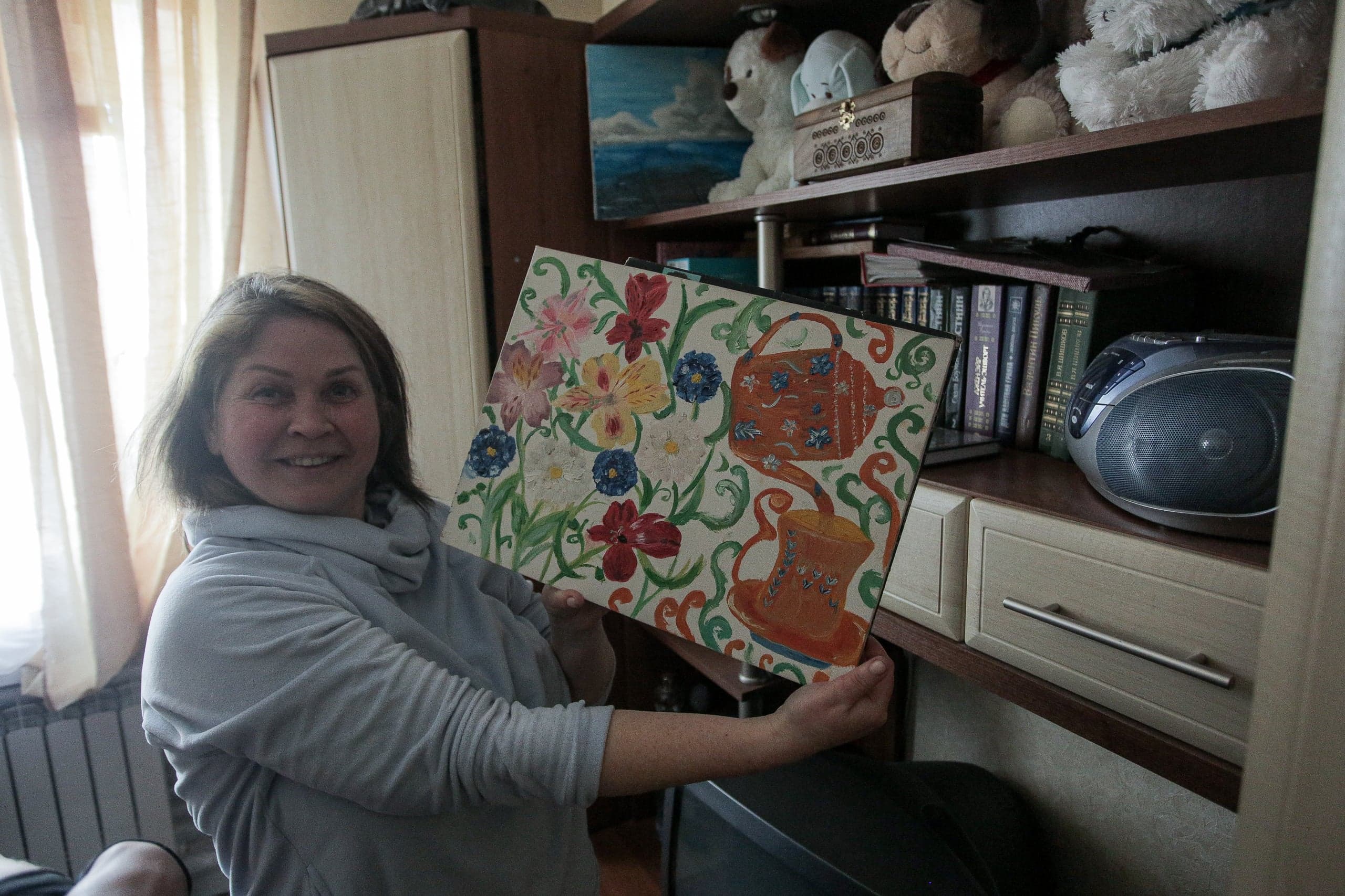 Улыбающаяся женщина демонстрирует картину с узором из ярких цветов и оранжевого чайника на фоне книжных полок.