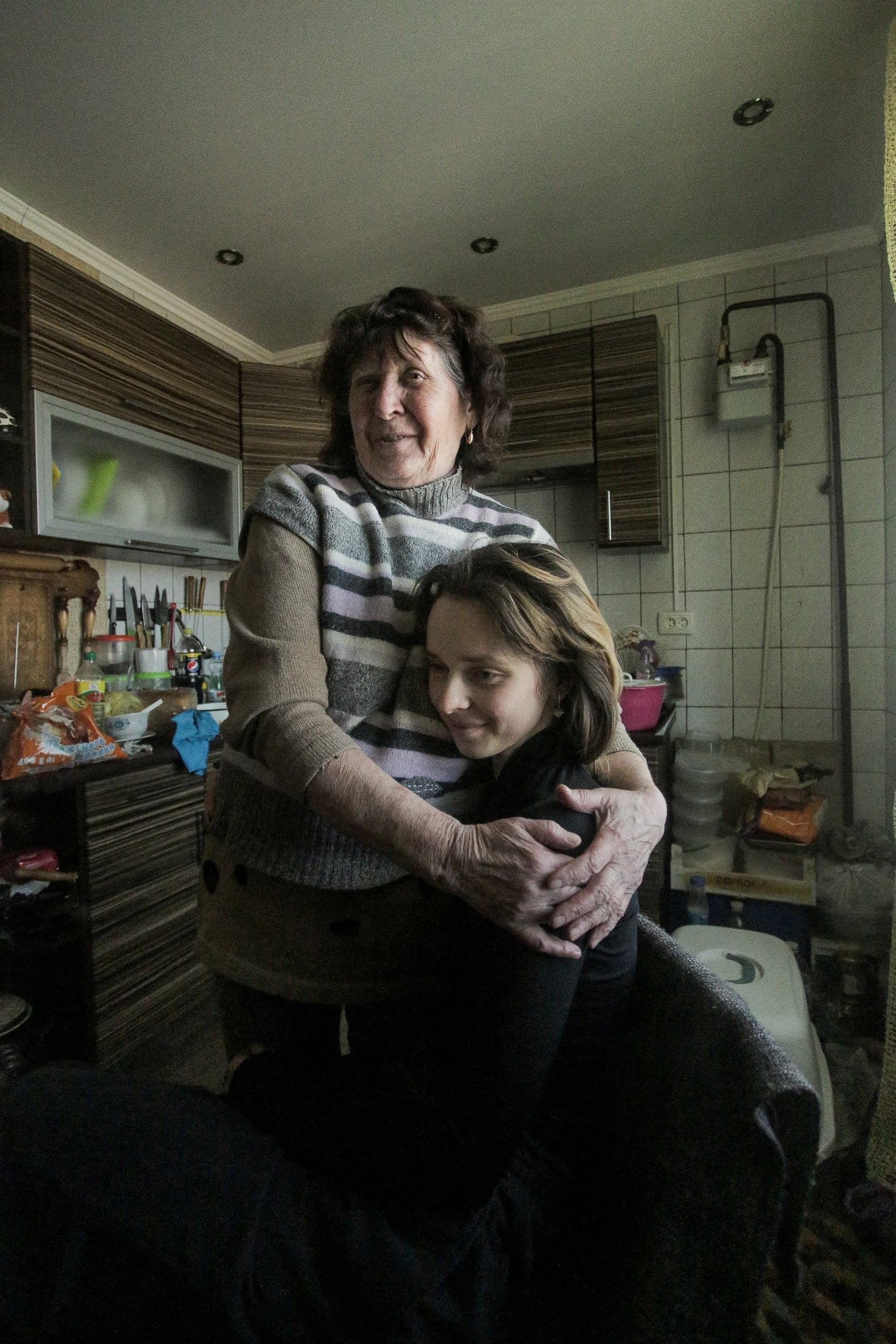 Пожилая женщина обнимает молодую женщину на захламлённой кухне; обе улыбаются.