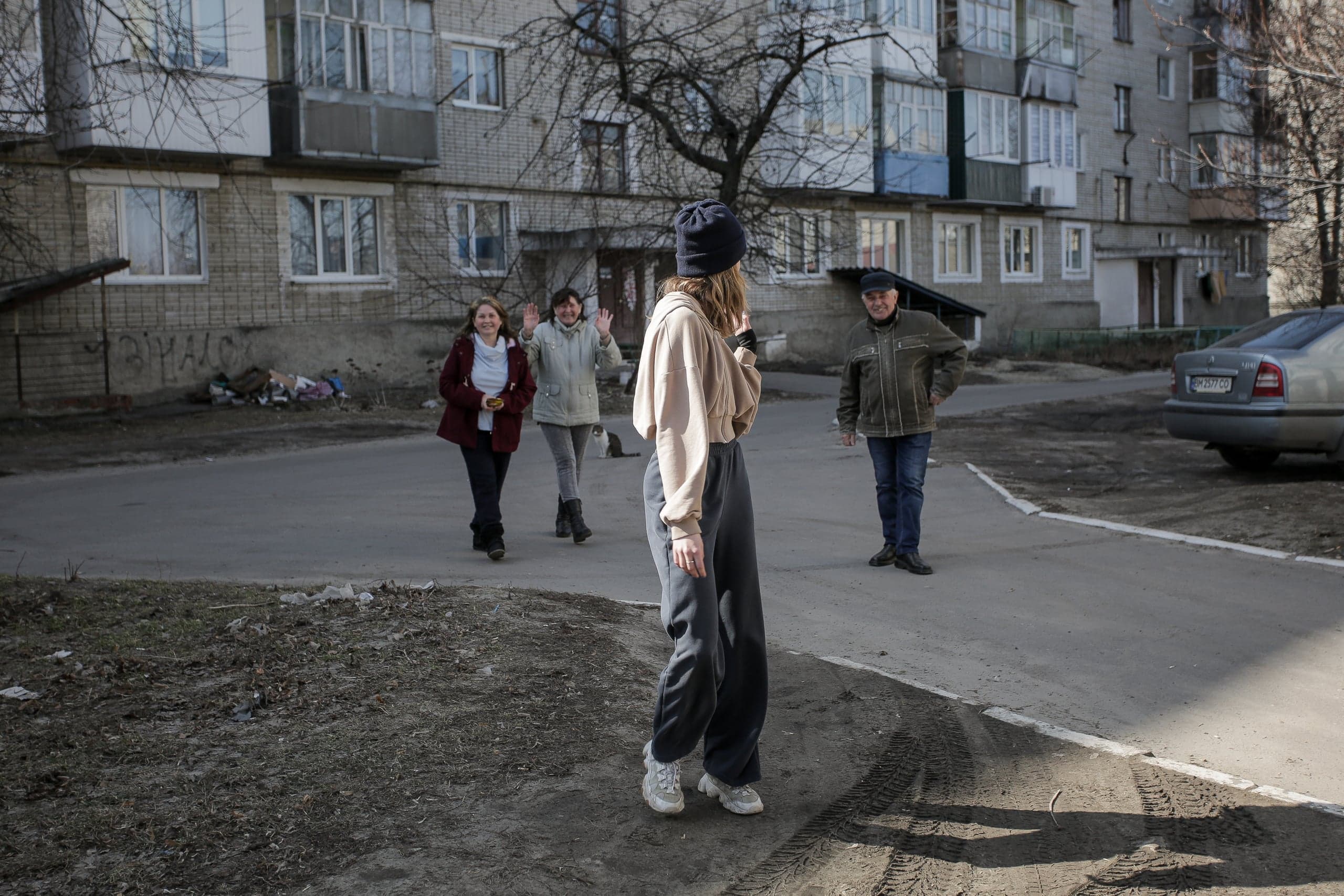 Люди приветствуют друг друга во дворе жилого дома. Молодой человек стоит спиной, двое взрослых машут ему.