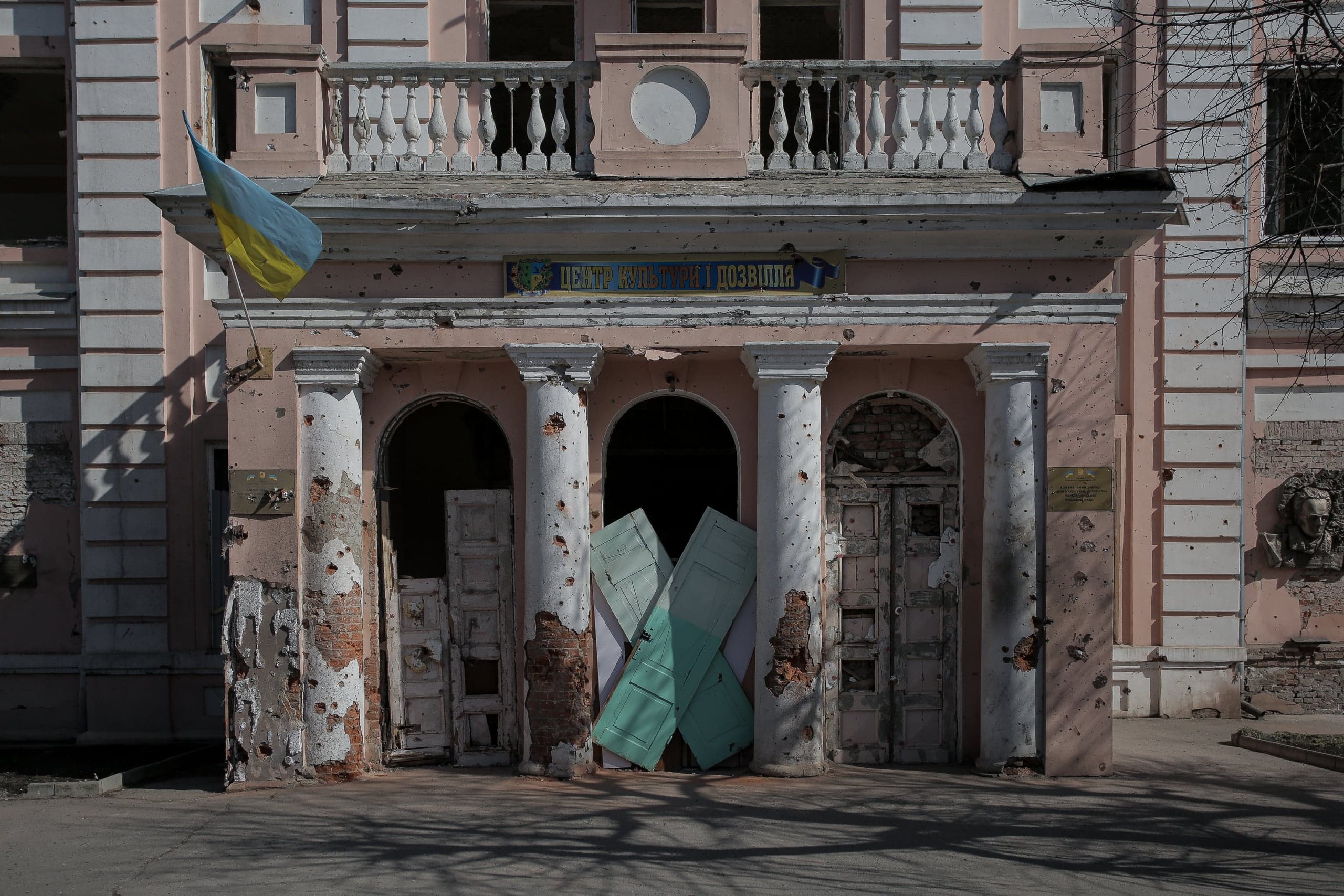 Поврежденный обстрелом фасад здания с колоннами и украинским флагом. Фасад усеян следами от пуль; над входом надпись: "ЦЕНТР КУЛЬТУРИ І ДОЗВІЛЛЯ".