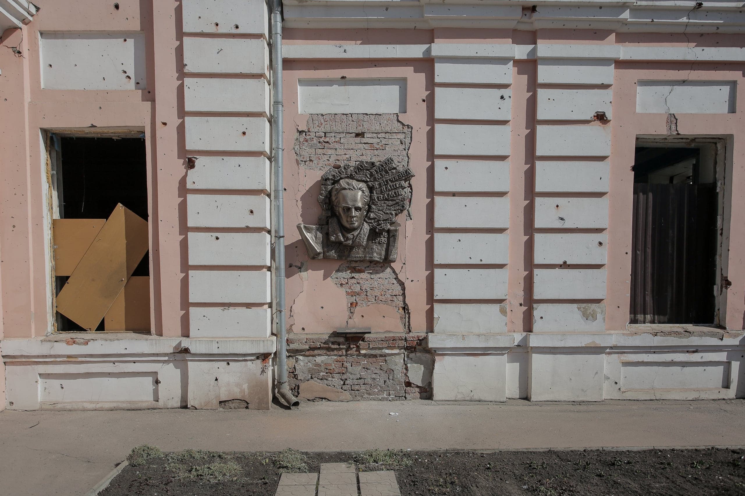 Фасад поврежденного розового здания, испещренный следами обстрела и пулевыми отверстиями. В центре – бронзовый барельеф мужчины, окруженный обвалившейся штукатуркой, обнажившей кирпичную кладку. Оконные проемы пустые.