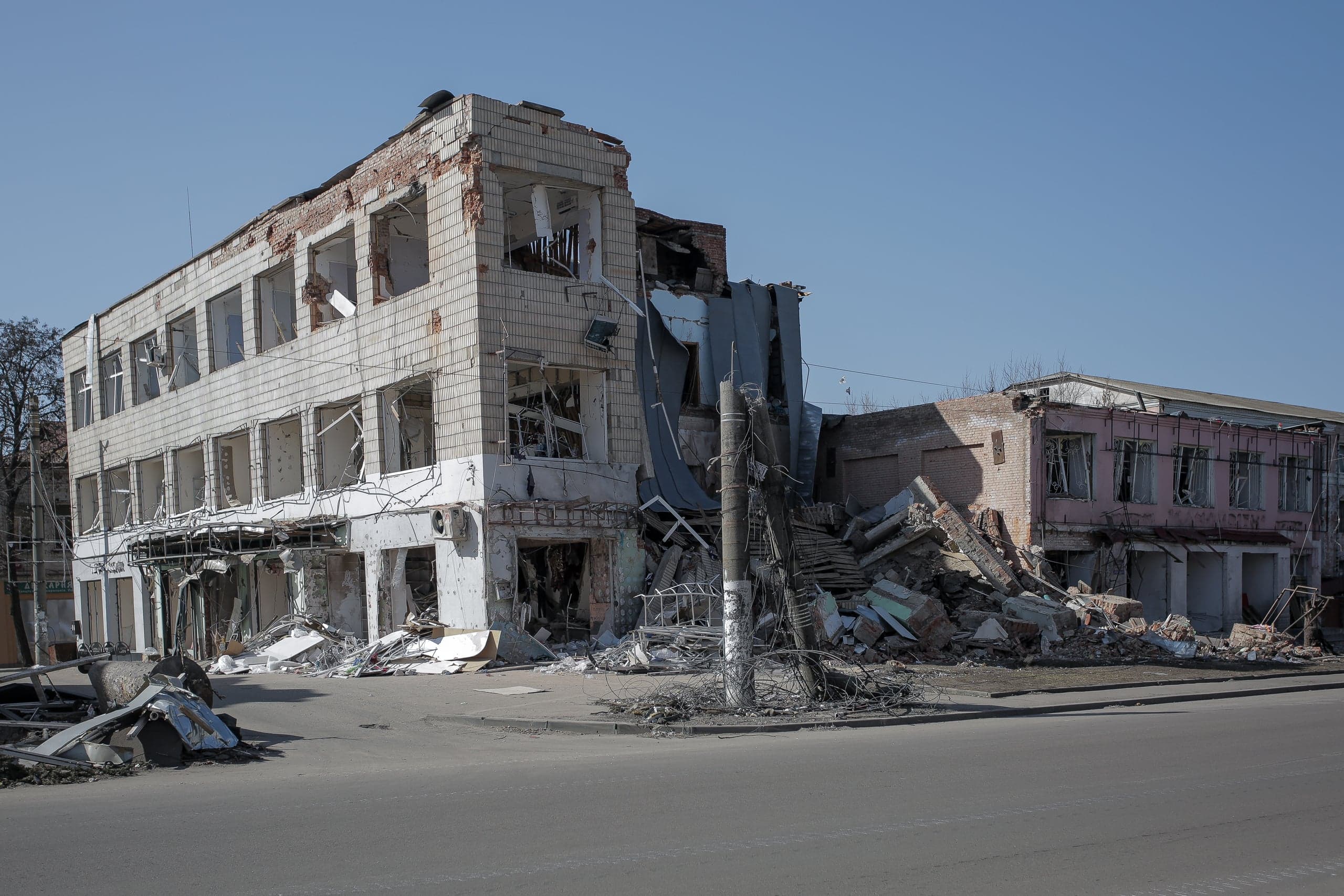 Разрушенные войной многоэтажное и прилегающее кирпичное здания на улице. Фасады и окна повреждены, повсюду обломки и руины.