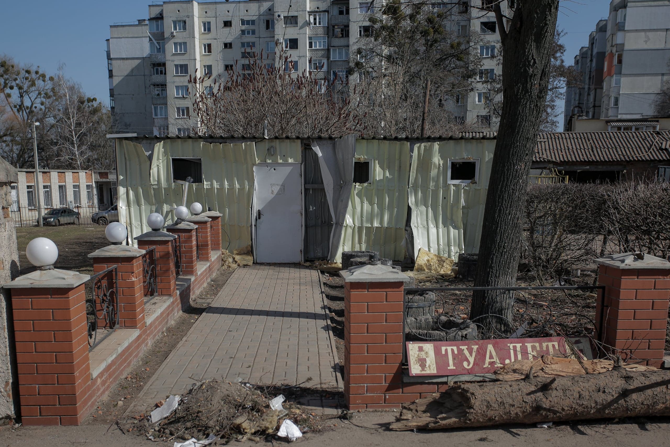 Поврежденная постройка из гофрированного металла. Вдоль дорожки — кирпичный забор с фонарями и знак "ТУАЛЕТ". На заднем плане — многоэтажный жилой дом.