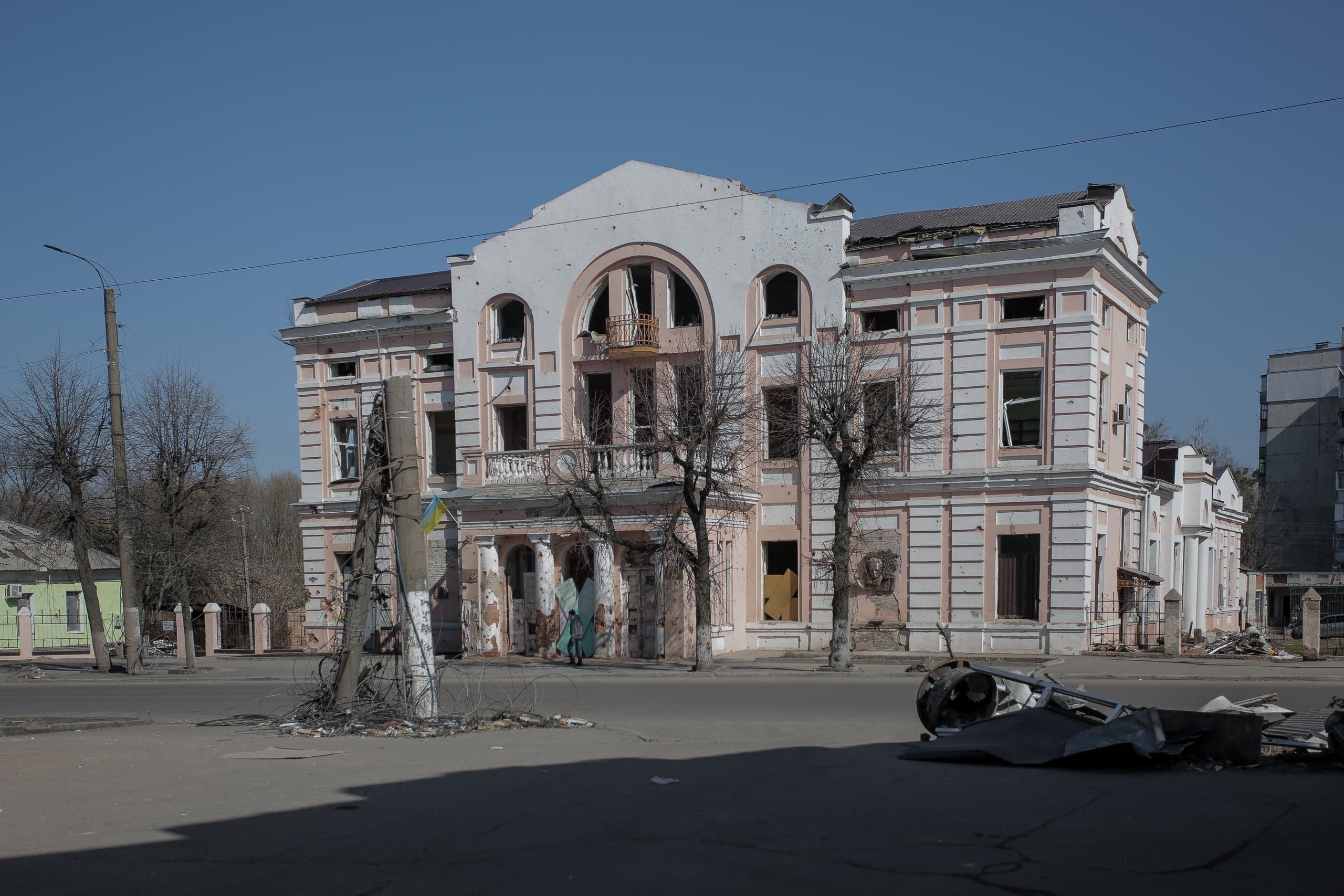 Разрушенное здание Дома культуры с выбитыми окнами и следами обстрела; на переднем плане обломки и поваленный столб.