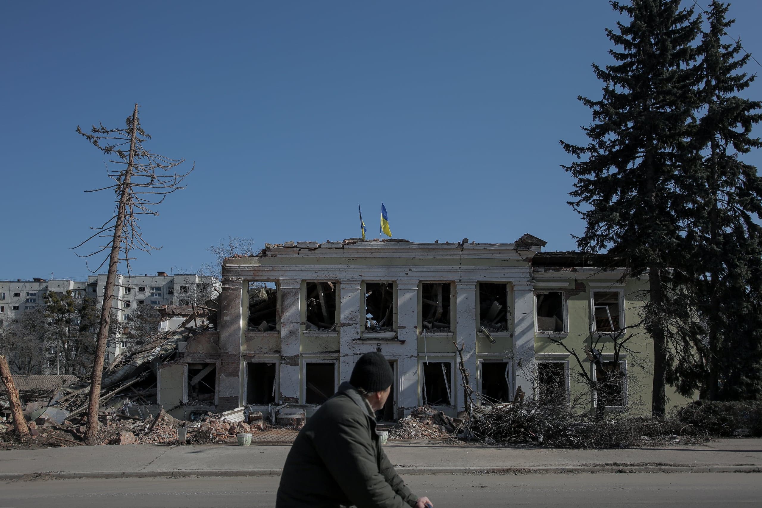 Разрушенное обстрелами здание с пустыми оконными проемами, на крыше которого развеваются два украинских флага. На переднем плане проходит человек.