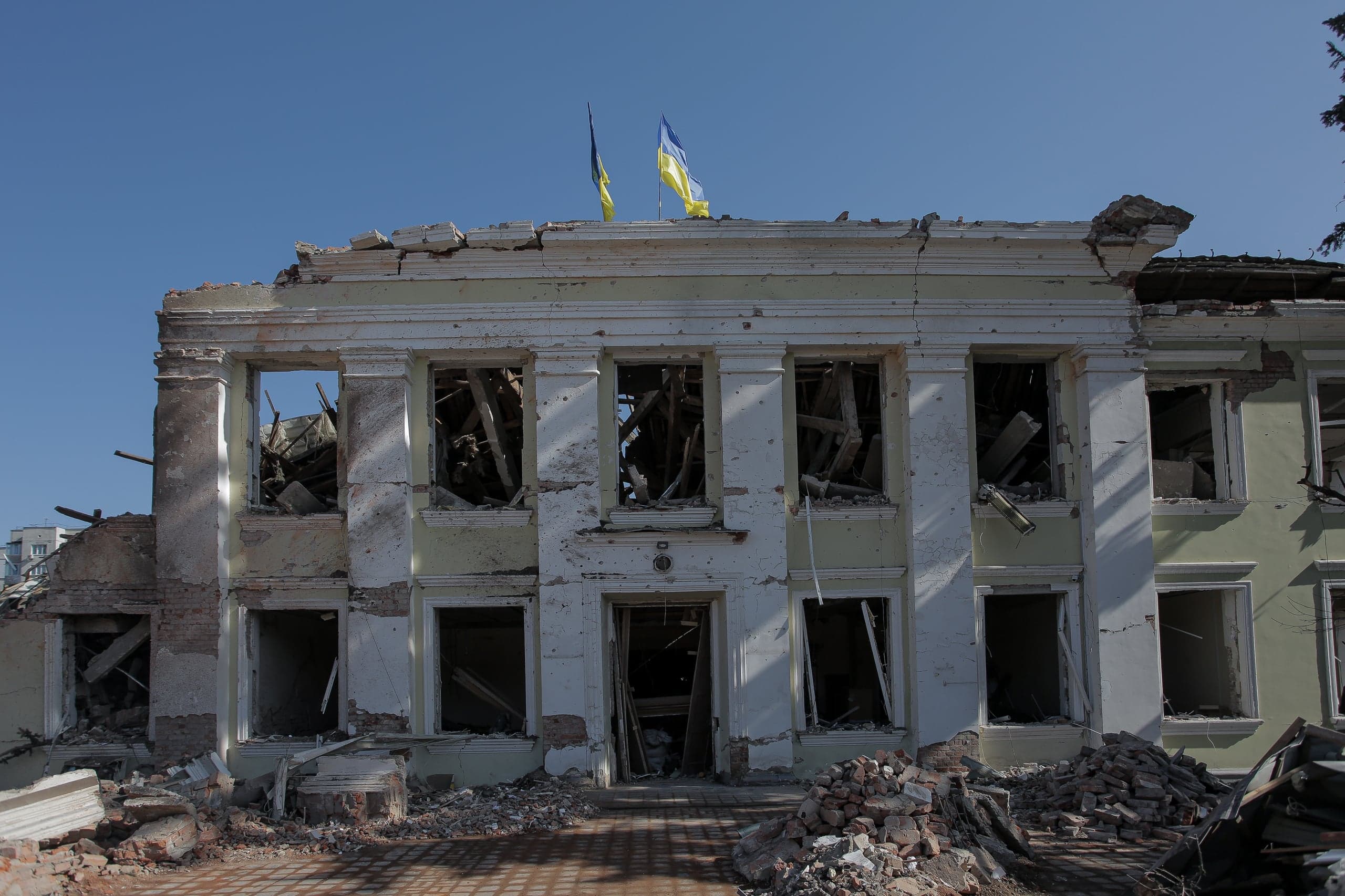 Разрушенное здание с двумя украинскими флагами на крыше. Фасад с выбитыми окнами, перед входом — груды обломков.
