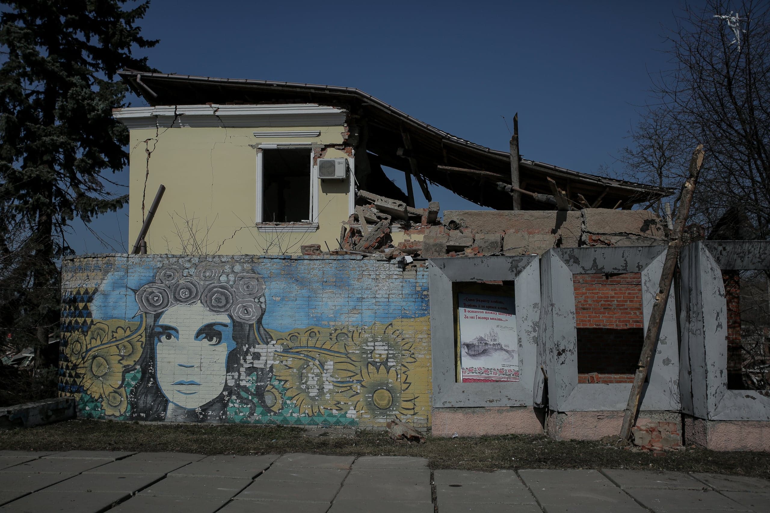 Разрушенное здание под голубым небом. Нижняя стена украшена большим граффити: женское лицо в венке среди сине-желтых подсолнухов. Видны обломки и обвалившаяся крыша.