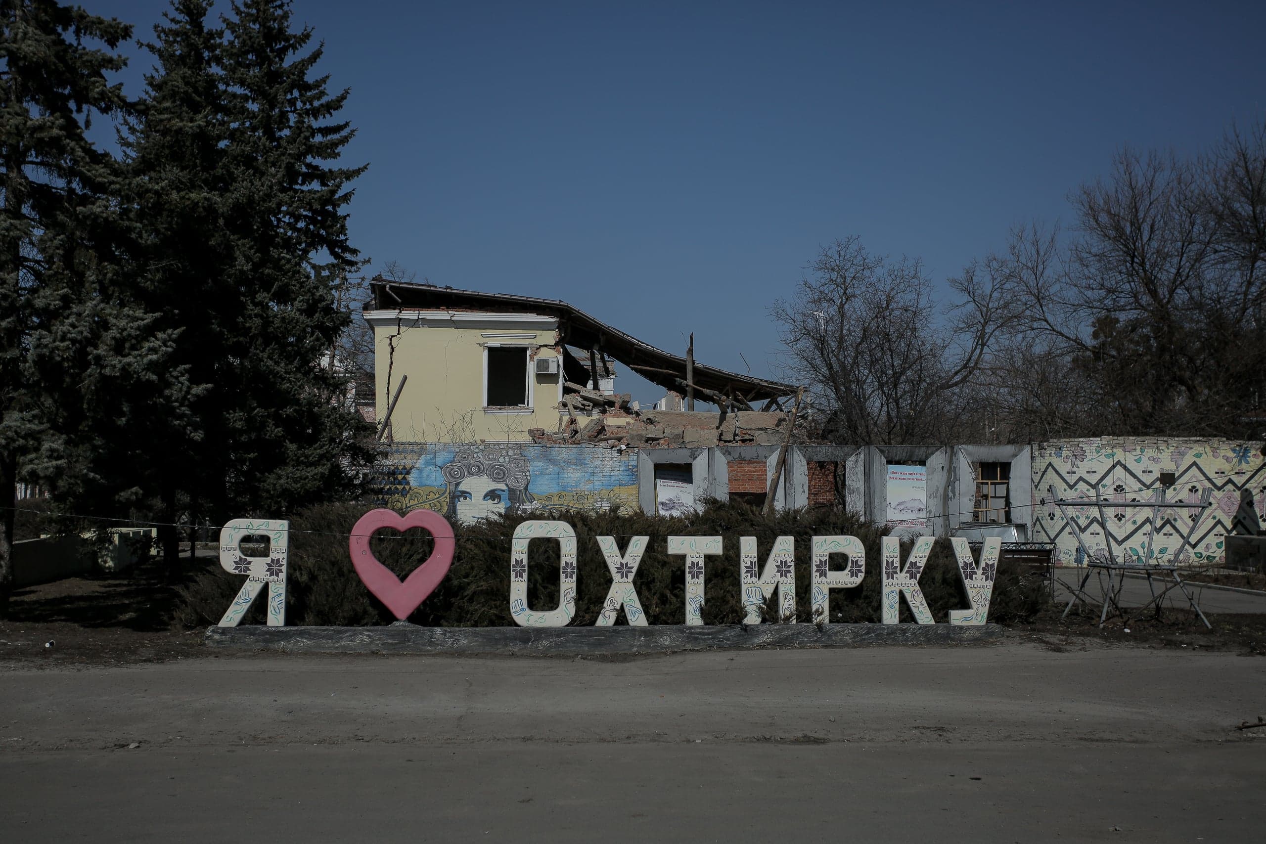 Надпись "Я ❤️ ОХТИРКУ" на фоне сильно поврежденного, частично разрушенного здания в городе Ахтырка.
