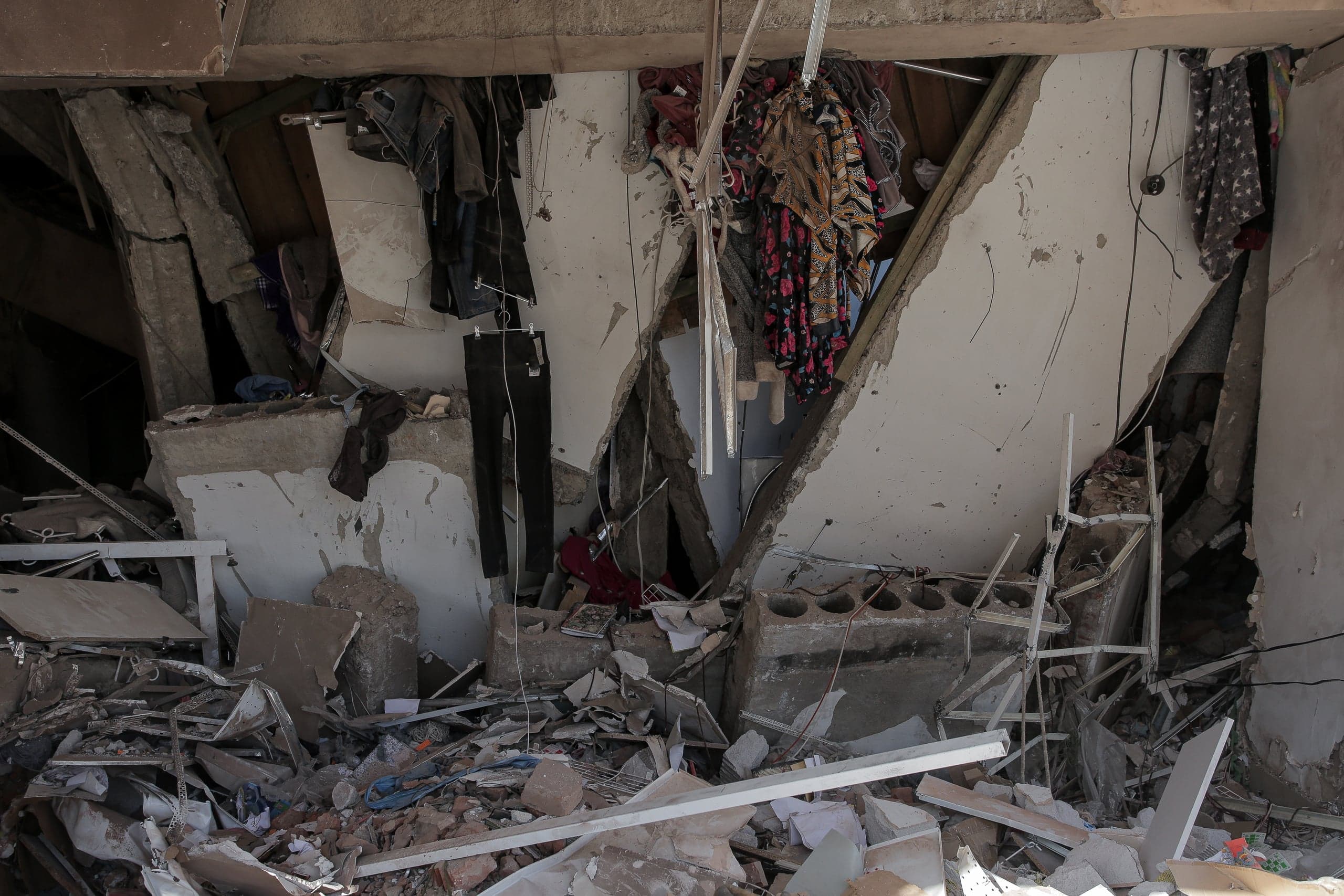 Интерьер разрушенного помещения, заваленный обломками бетона и кирпича. На заднем плане на уцелевших фрагментах стен висит одежда.