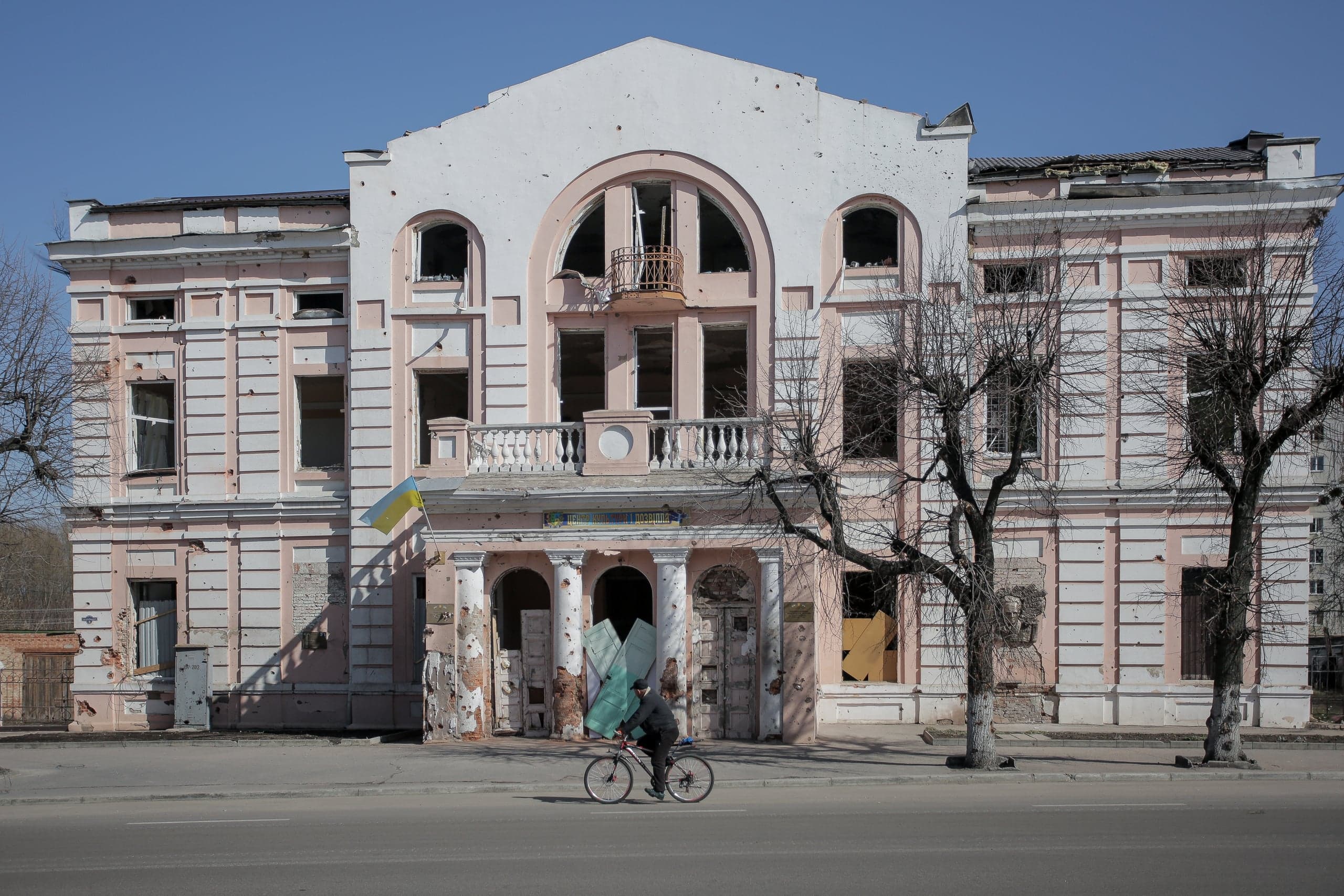 Сильно поврежденное снарядами розово-белое здание с выбитыми окнами. Человек на велосипеде проезжает мимо фасада, на котором виден украинский флаг.