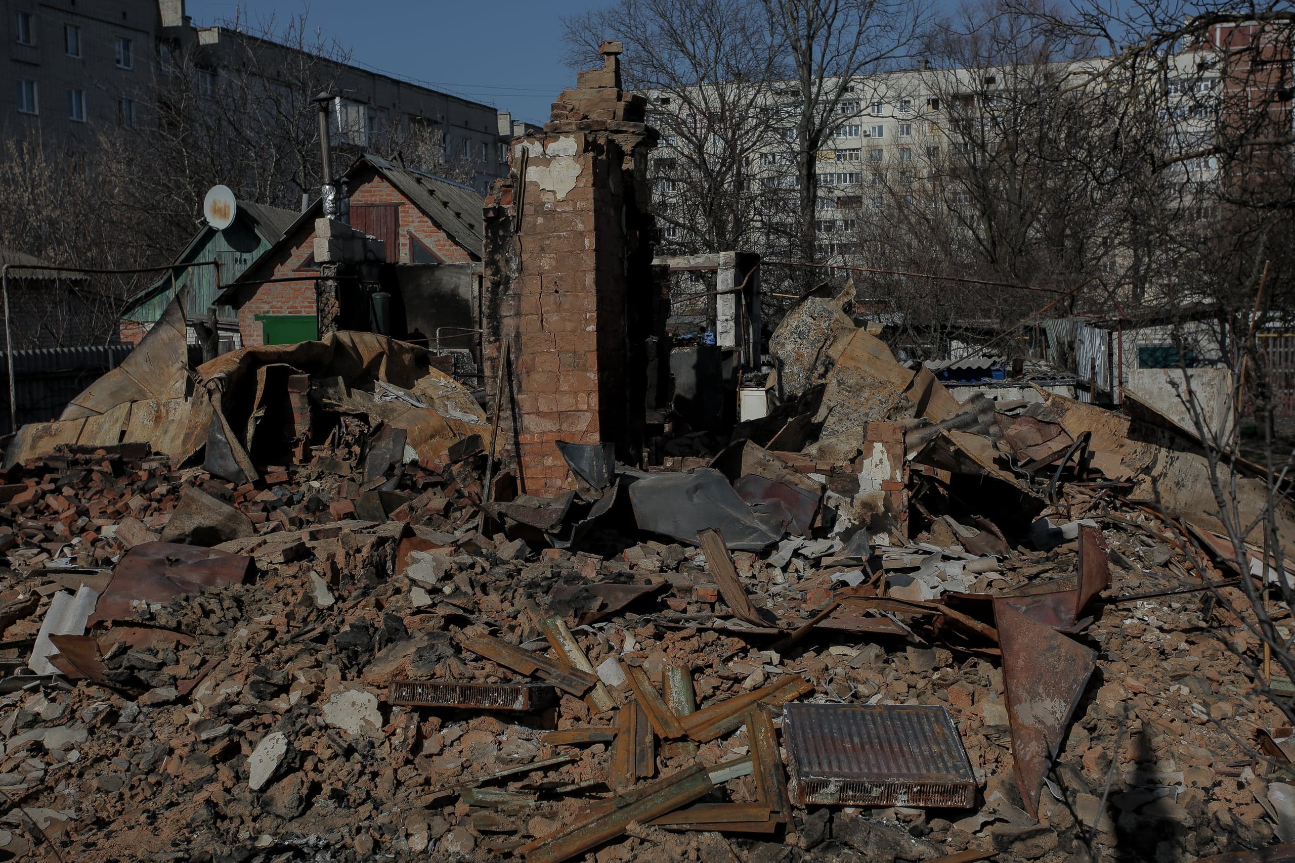 Руины частного дома с остатками кирпичной стены и горой обломков на переднем плане, на фоне жилых многоэтажек.