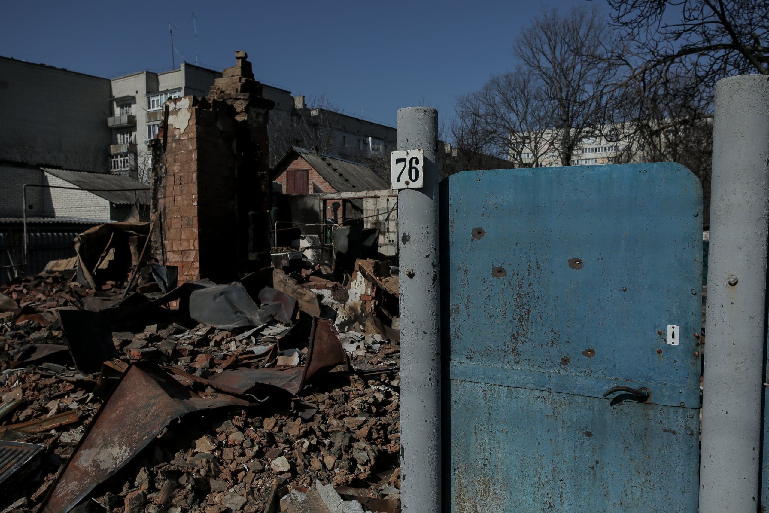 Разрушенный обстрелом дом, остатки кирпичной стены и обломки на фоне синих ворот с табличкой "76" на столбе.