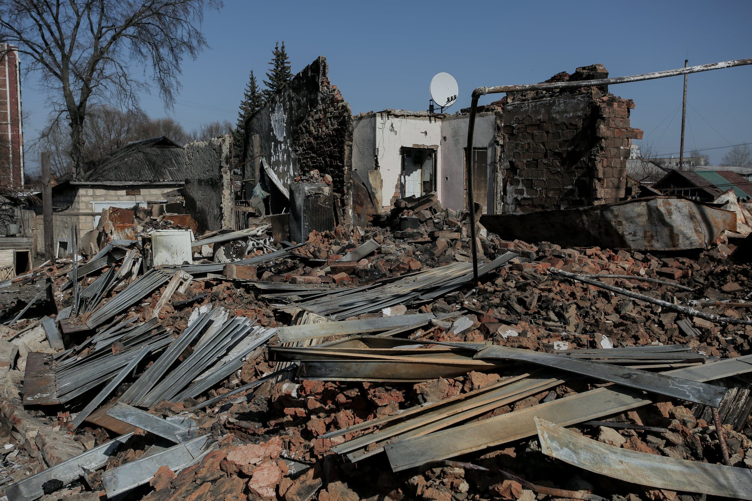 Разрушенный жилой дом, заваленный обломками кирпича и металлическим мусором. На фоне — обгоревшие остатки стен со спутниковой антенной.