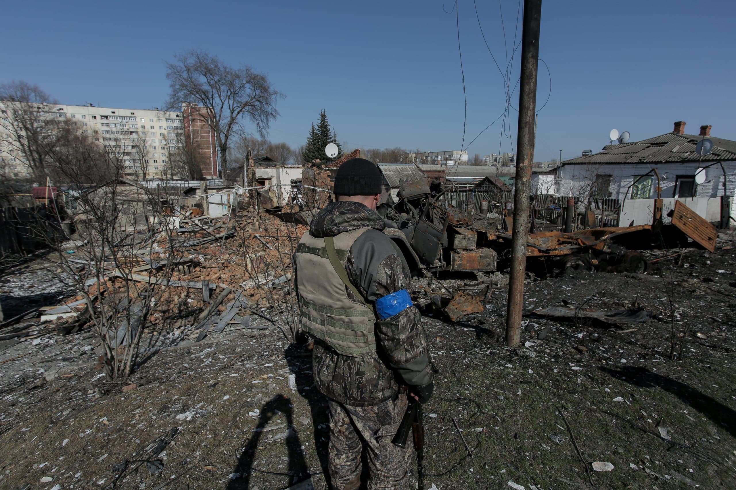 Военнослужащий с синей повязкой осматривает руины частных домов и сгоревшую технику в разрушенном городском квартале. На заднем плане — целые многоэтажные здания.