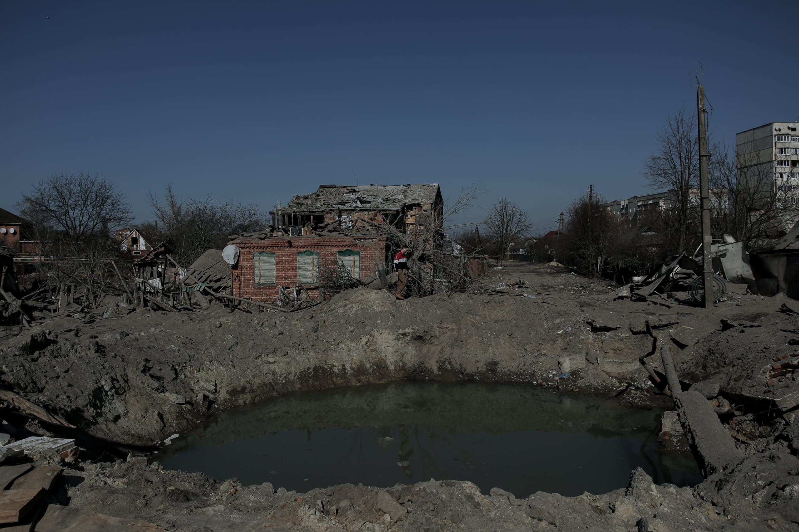 Большая, заполненная водой воронка перед сильно разрушенным кирпичным домом посреди руин городского района.