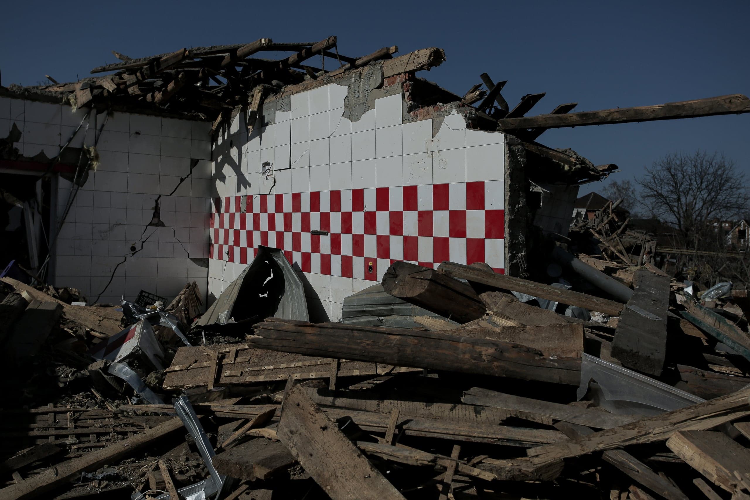 Разрушенное здание с частично уцелевшей стеной из белой плитки и красно-белой клетчатой полосой. На переднем плане — горы обломков и деревянных балок.
