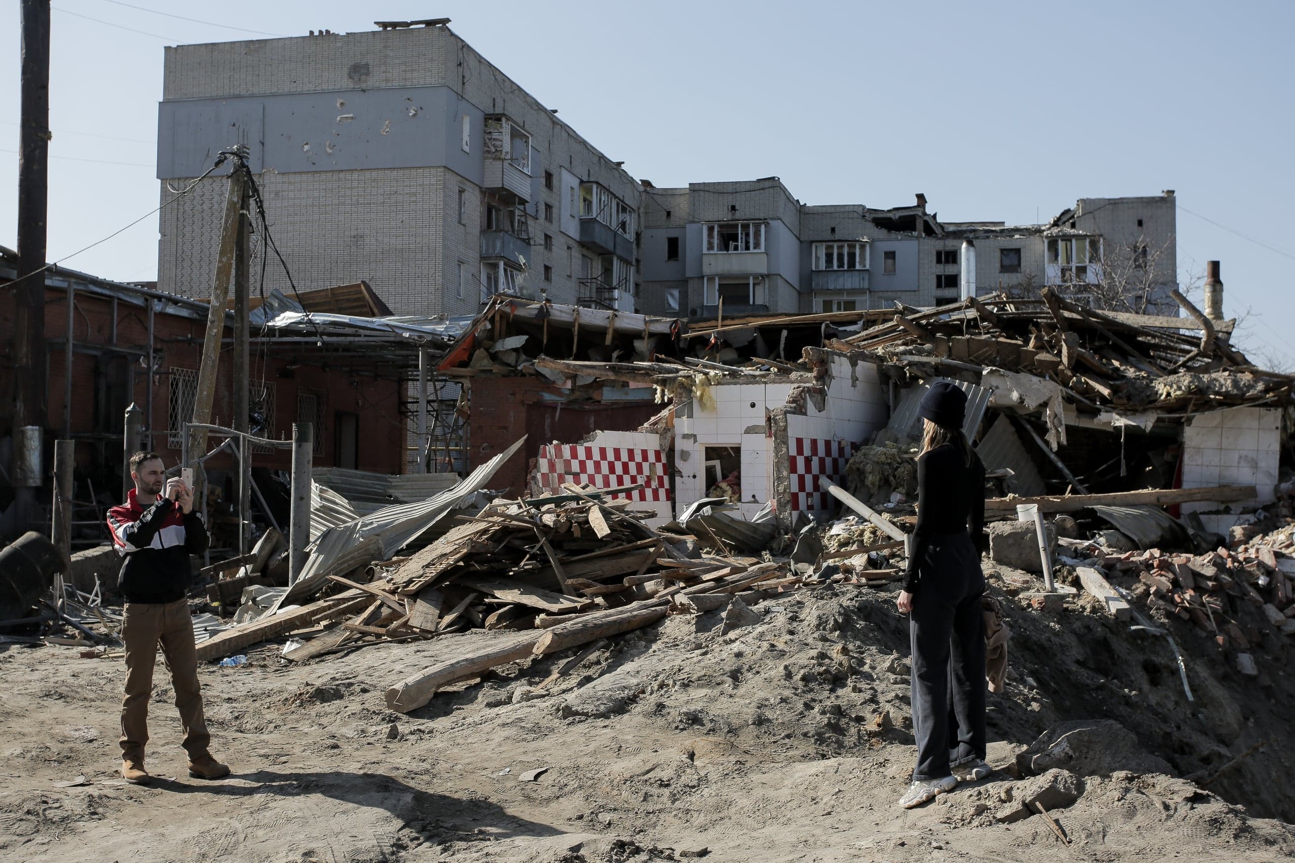 Мужчина и женщина стоят перед грудой обломков разрушенного здания, на фоне многоэтажный дом с повреждениями от обстрелов.