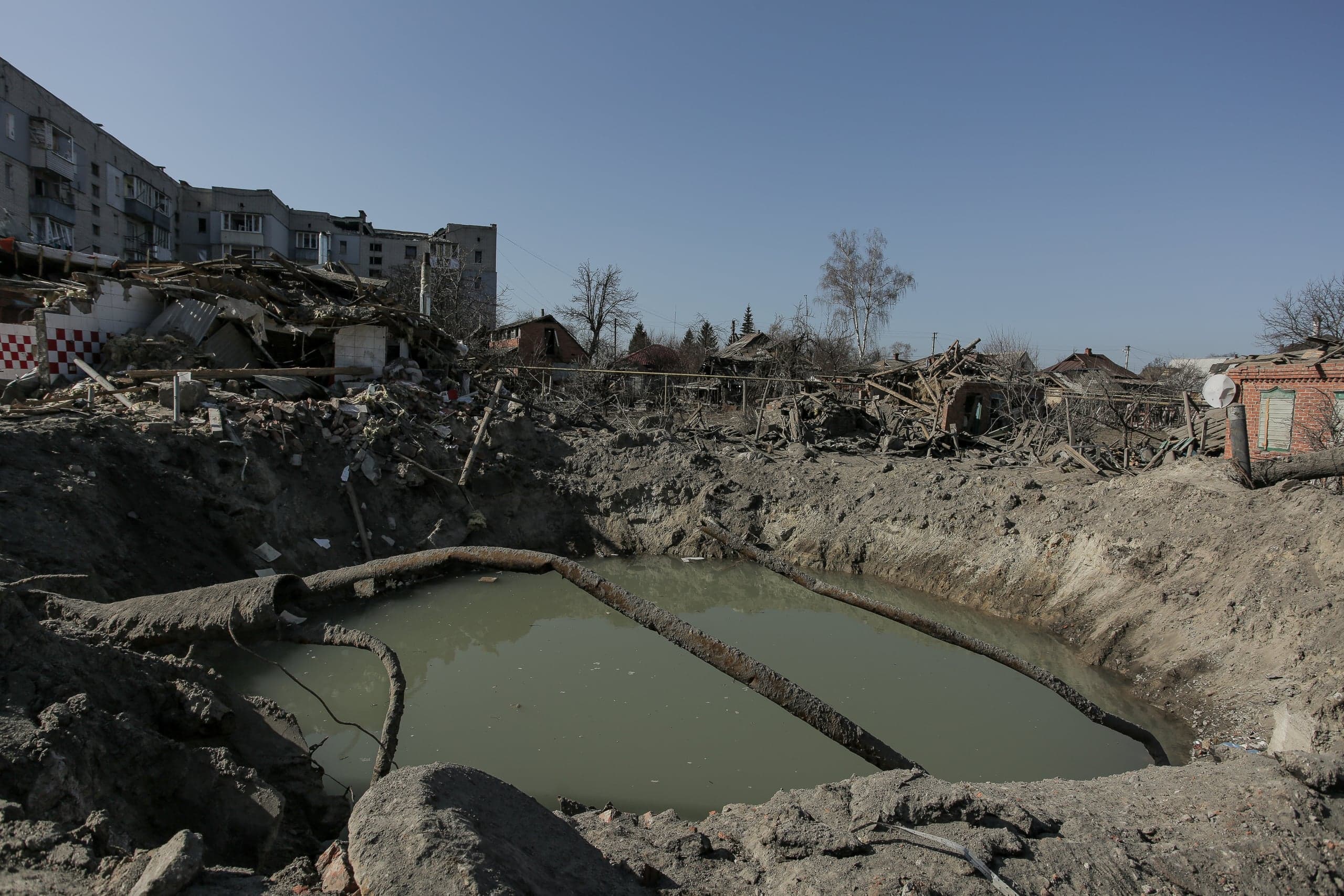 Крупная воронка от взрыва, заполненная водой, на фоне разрушенных многоэтажных и частных жилых зданий.