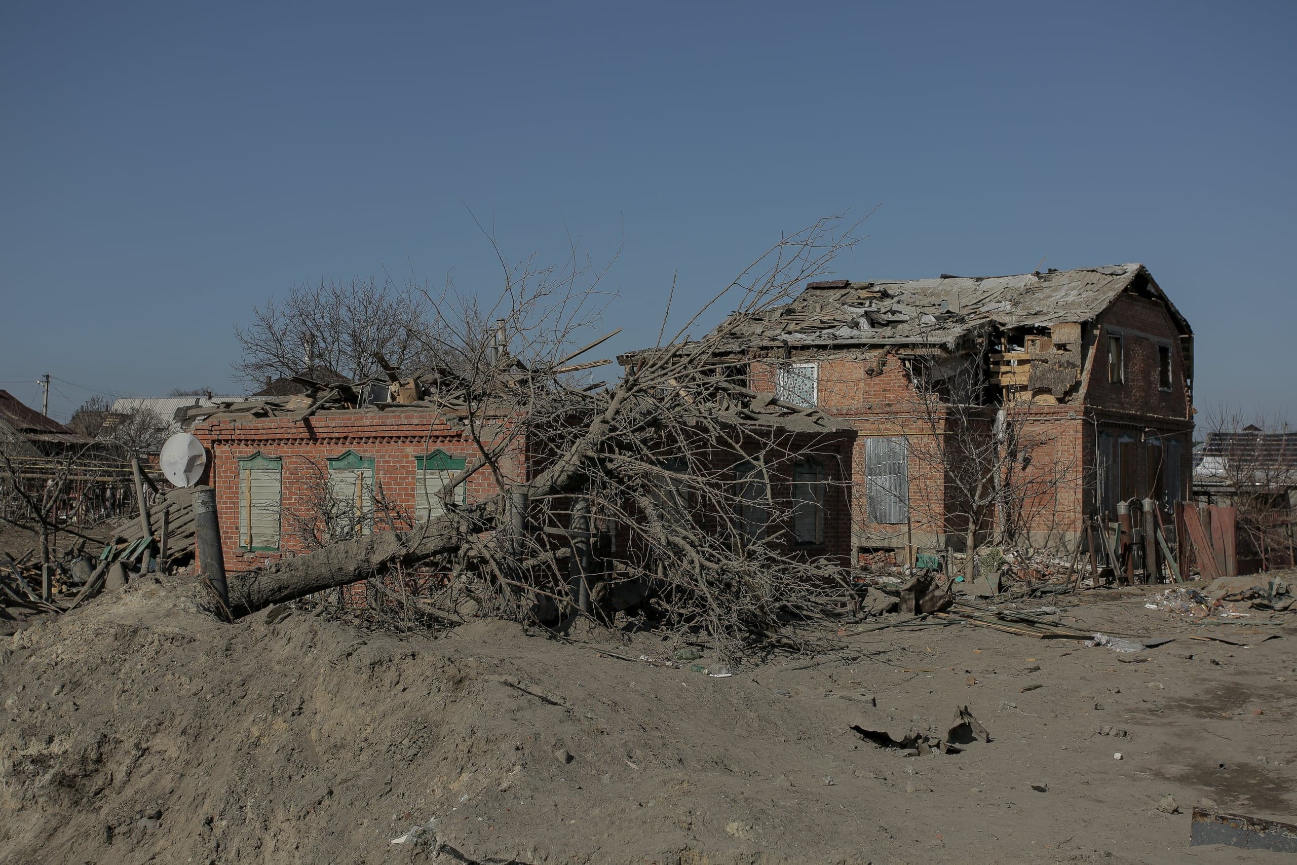 Разрушенные обстрелами кирпичные дома под ясным небом, заваленные обломками и поваленными деревьями.