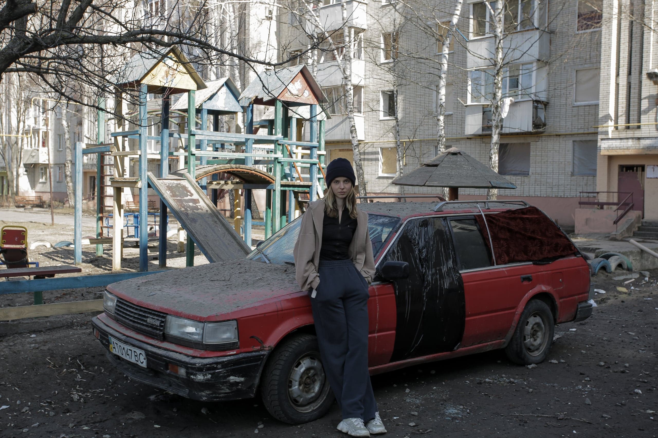 Женщина у грязного красного универсала, окна которого заклеены пленкой и тканью, на фоне заброшенной детской площадки и жилого дома. Номер "AT 0147 BC".