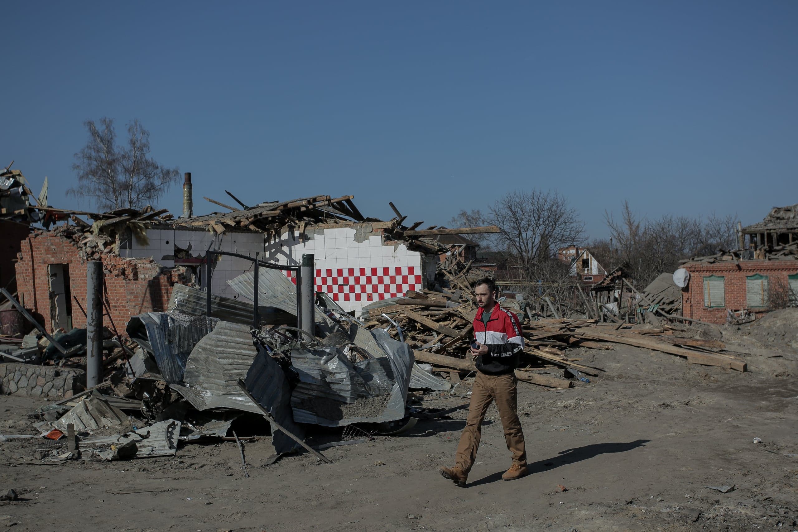Мужчина идет через руины разрушенного жилого района. На заднем плане — обломки домов и стена с красно-белым клетчатым узором.