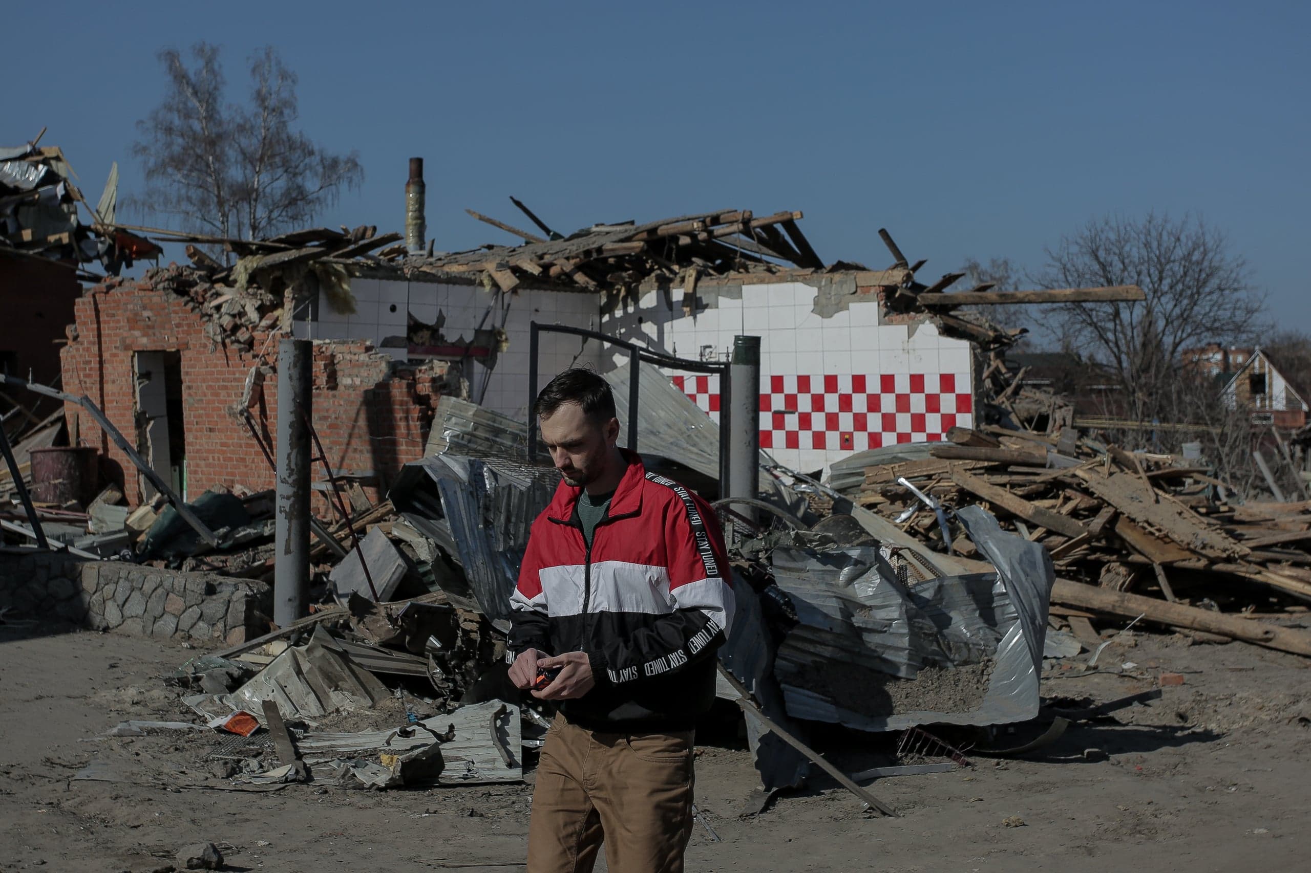 Мужчина стоит посреди руин и обломков разрушенного здания; виден фрагмент стены с красно-белым клетчатым узором.
