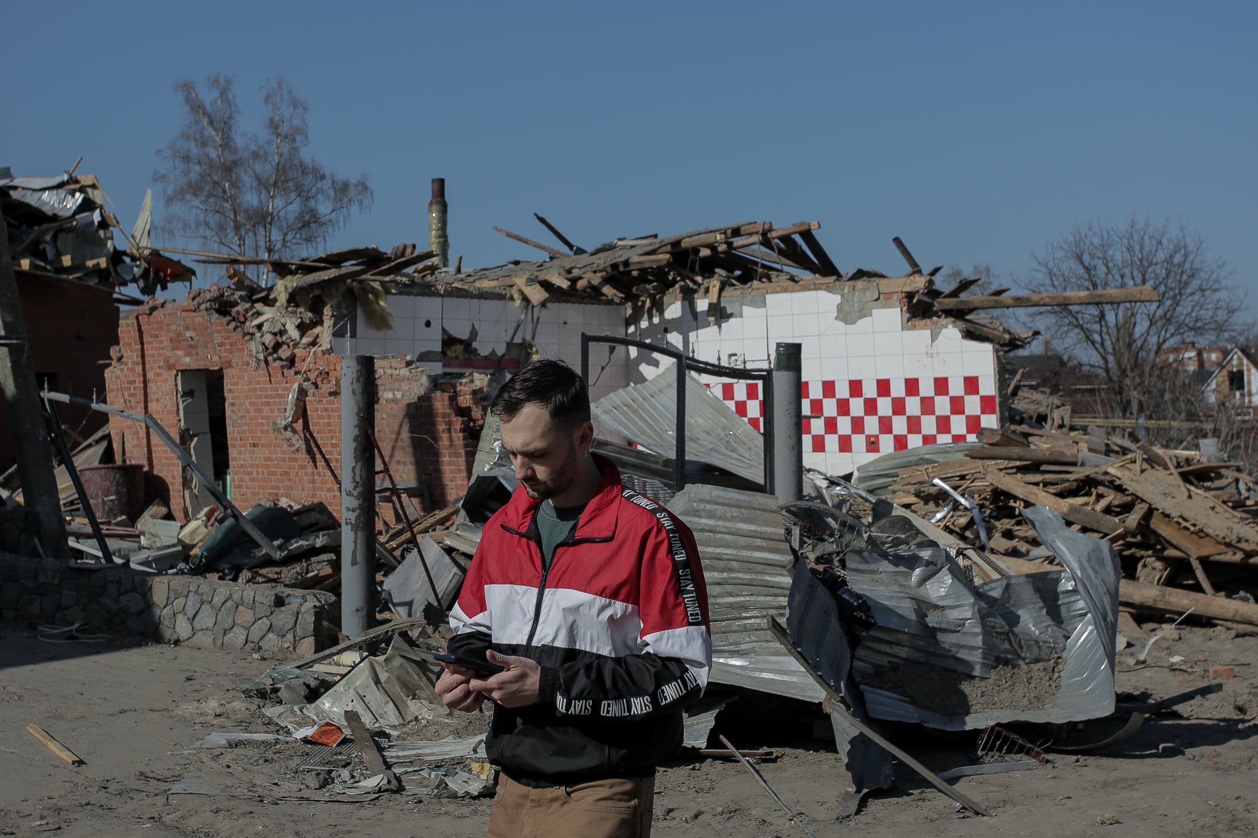 Мужчина смотрит в телефон, стоя среди обломков разрушенных зданий, включая стену с красно-белым клетчатым узором.