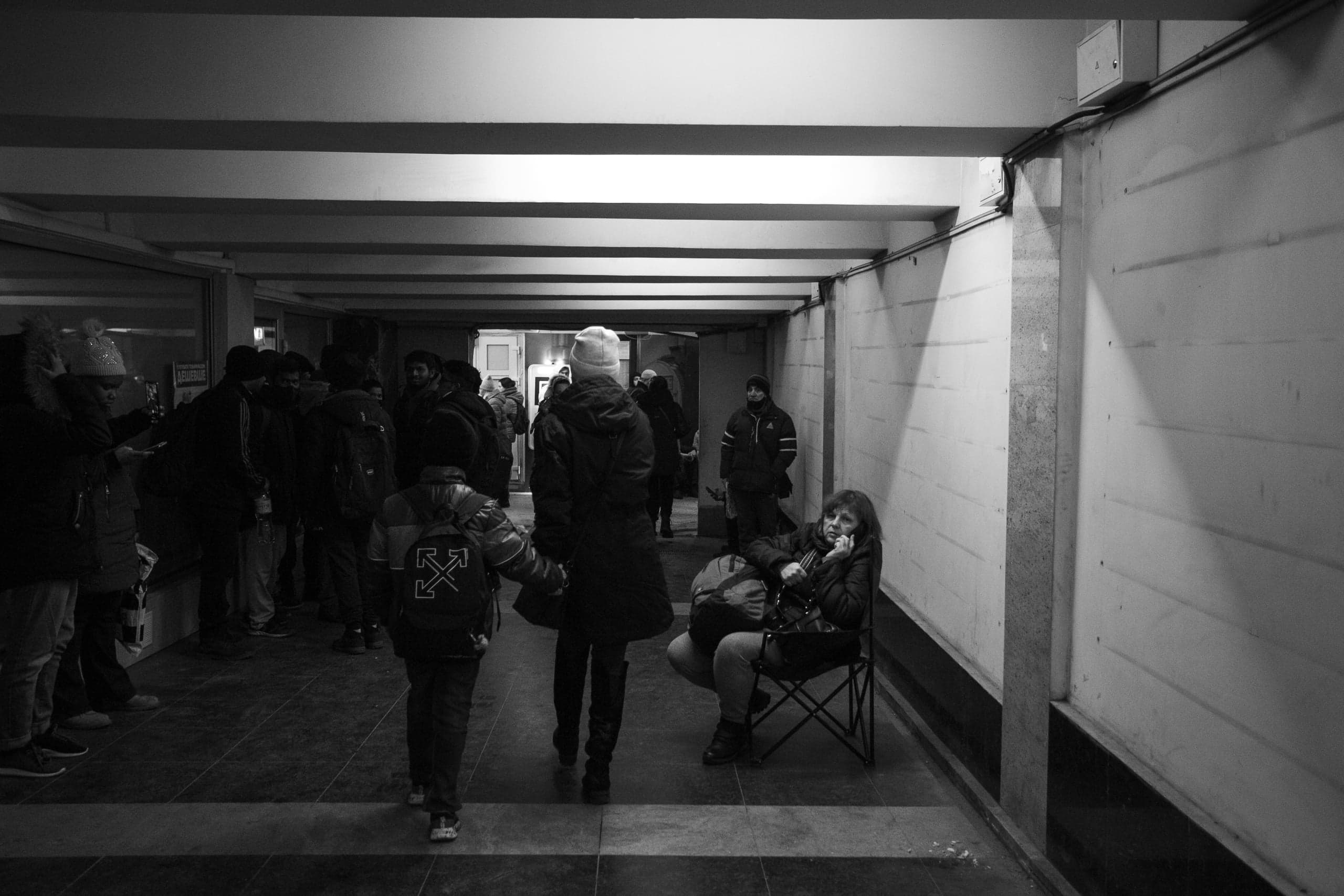 Черно-белое фото: толпа людей в темном подземном переходе или убежище. Справа женщина сидит на складном стуле и разговаривает по телефону.