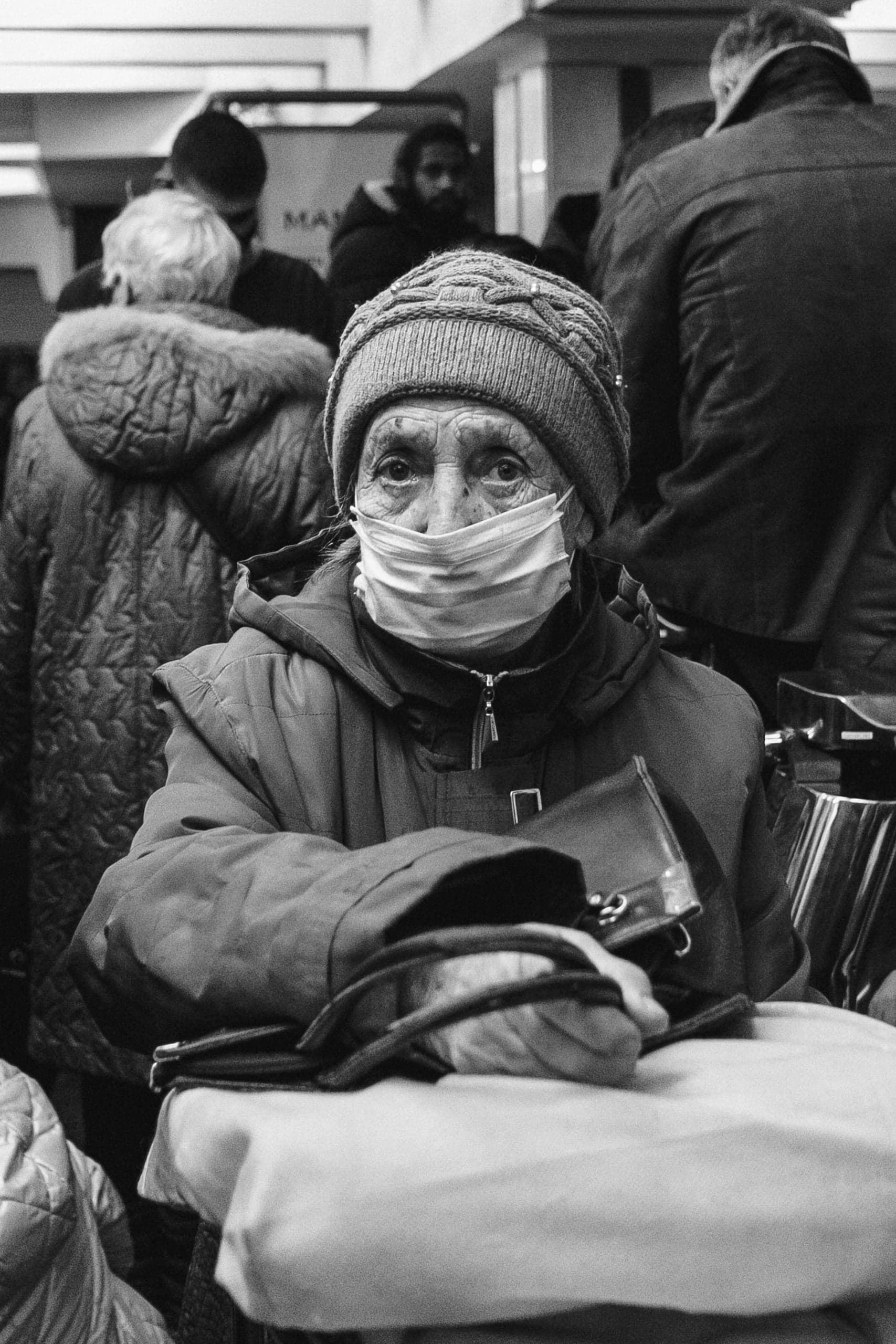 Пожилой человек в вязаной шапке и медицинской маске крупным планом на фоне толпы. Черно-белое фото.