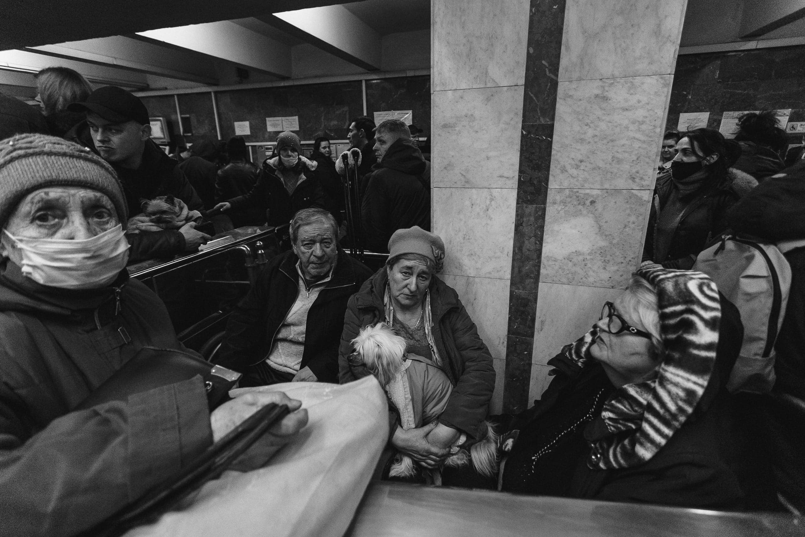 Черно-белое фото: пожилые люди укрываются в метро. Женщина в маске и вязаной шапке на переднем плане. Рядом сидит пара, женщина держит маленькую собаку.