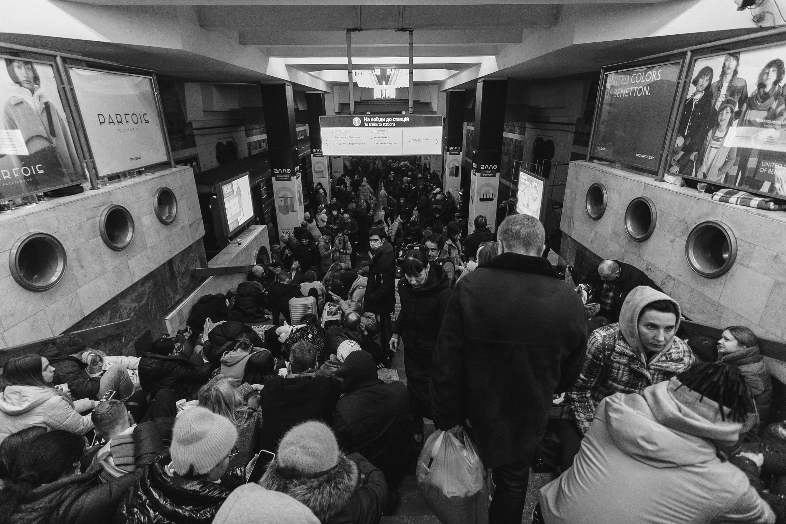 Черно-белое фото: толпа людей сидит в вестибюле метро, используемом как убежище. Рекламные постеры ("PARFOIS", "UNITED COLORS OF BENETTON") и табло "На поїзди до станції / To trains to station".