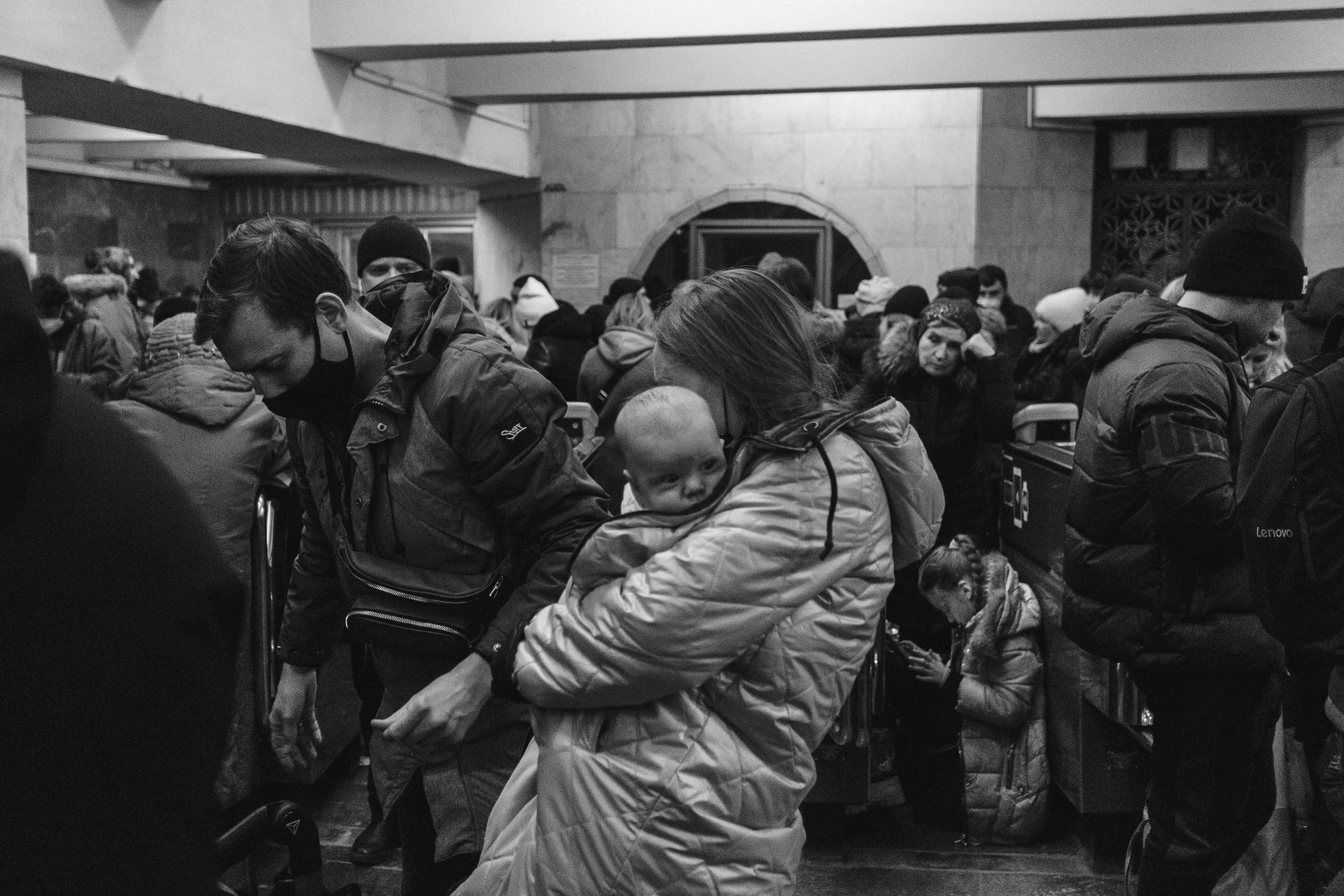 Черно-белое фото: толпа людей в помещении (вероятно, метро). Женщина держит на руках младенца, рядом стоит мужчина в маске.