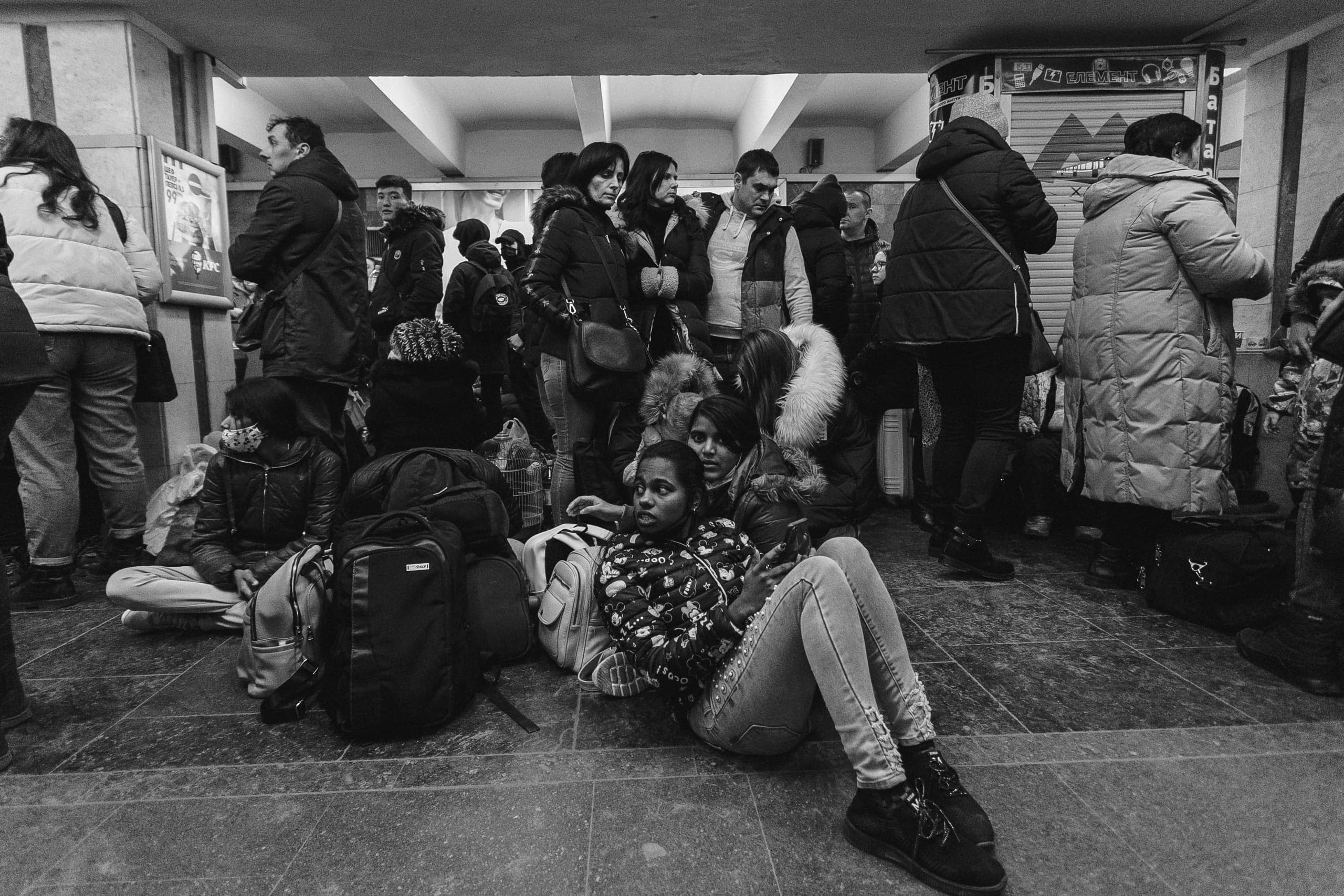 Черно-белое фото скопления людей и багажа в помещении. Женщины, сидящие на полу, окружены толпой.