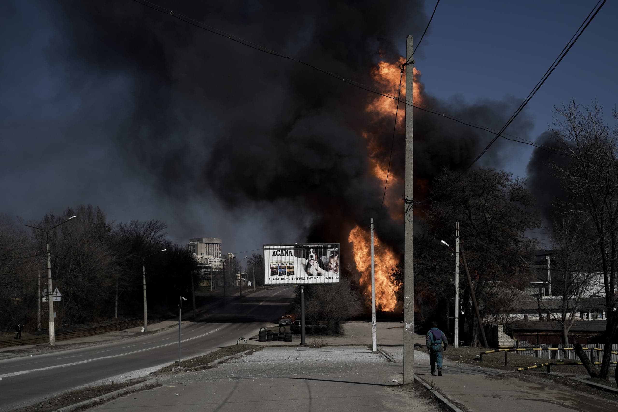 Уличная сцена, где огромное оранжевое пламя и столб черного дыма поднимаются в небо, заслоняя городские постройки. Пешеход в спецодежде идет по тротуару вдаль, мимо рекламного щита и бетонного столба, на фоне пожара.