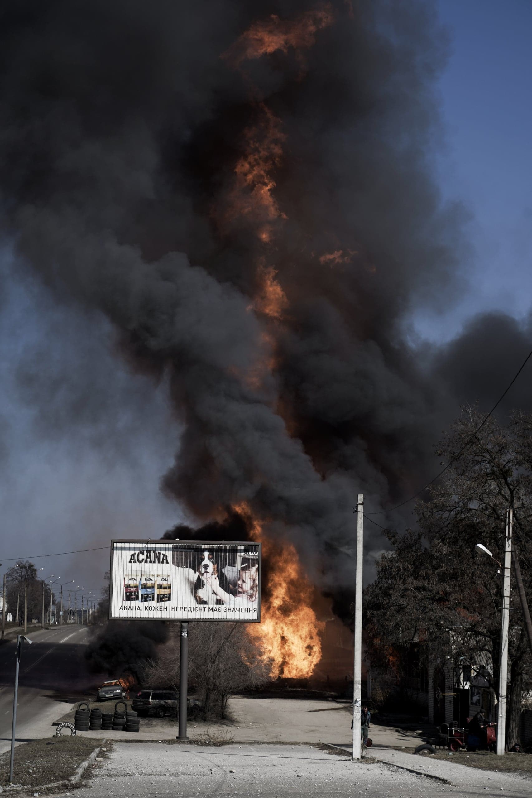 Уличный рекламный щит ACANA на фоне огромного столба черного дыма и оранжевого пламени, поднимающегося от масштабного пожара. Слева видны горящие автомобили и покрышки.