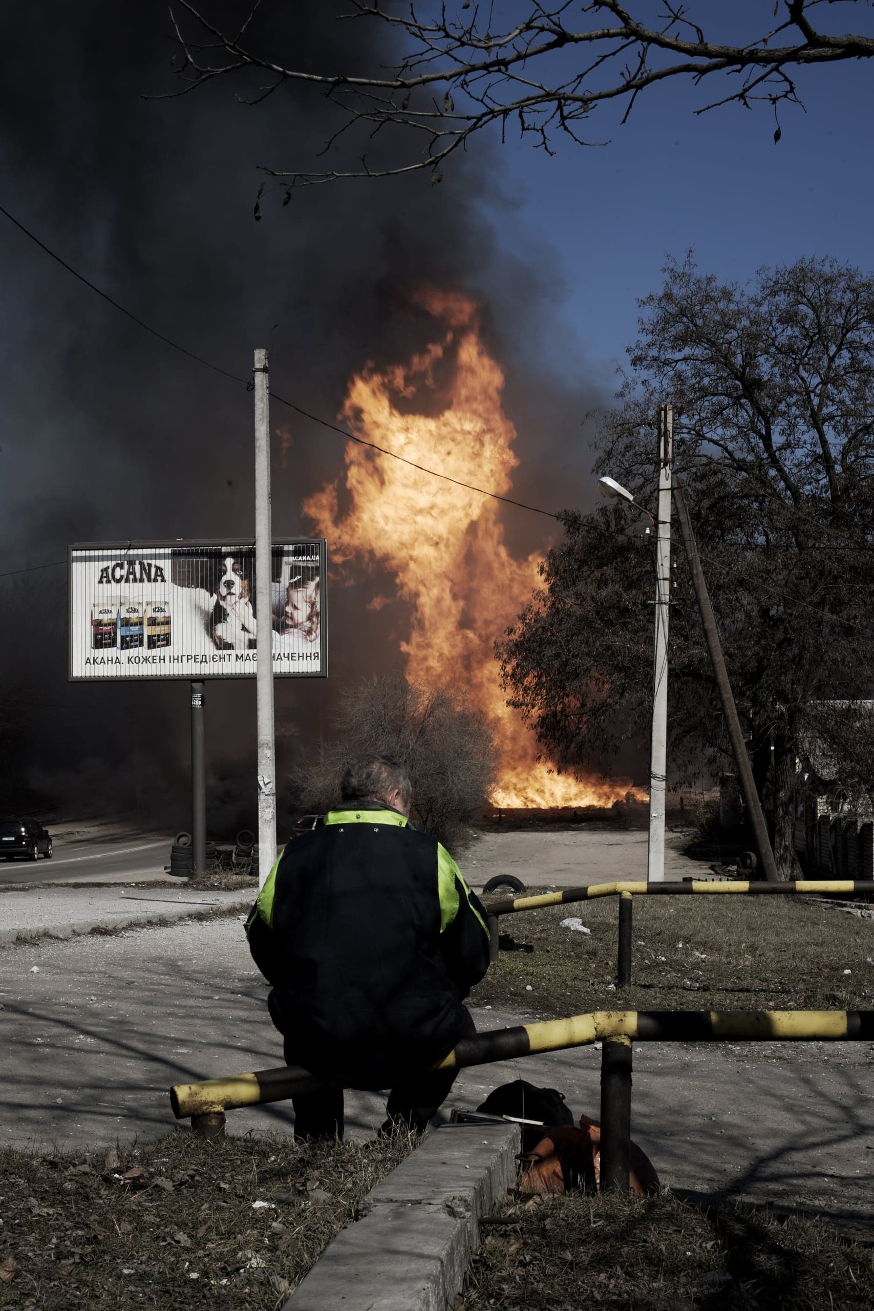 Человек в куртке, сидящий спиной к камере, наблюдает за огромным оранжевым пламенем и столбом черного дыма, которые поднимаются в небо за рекламным щитом с кормом для собак.