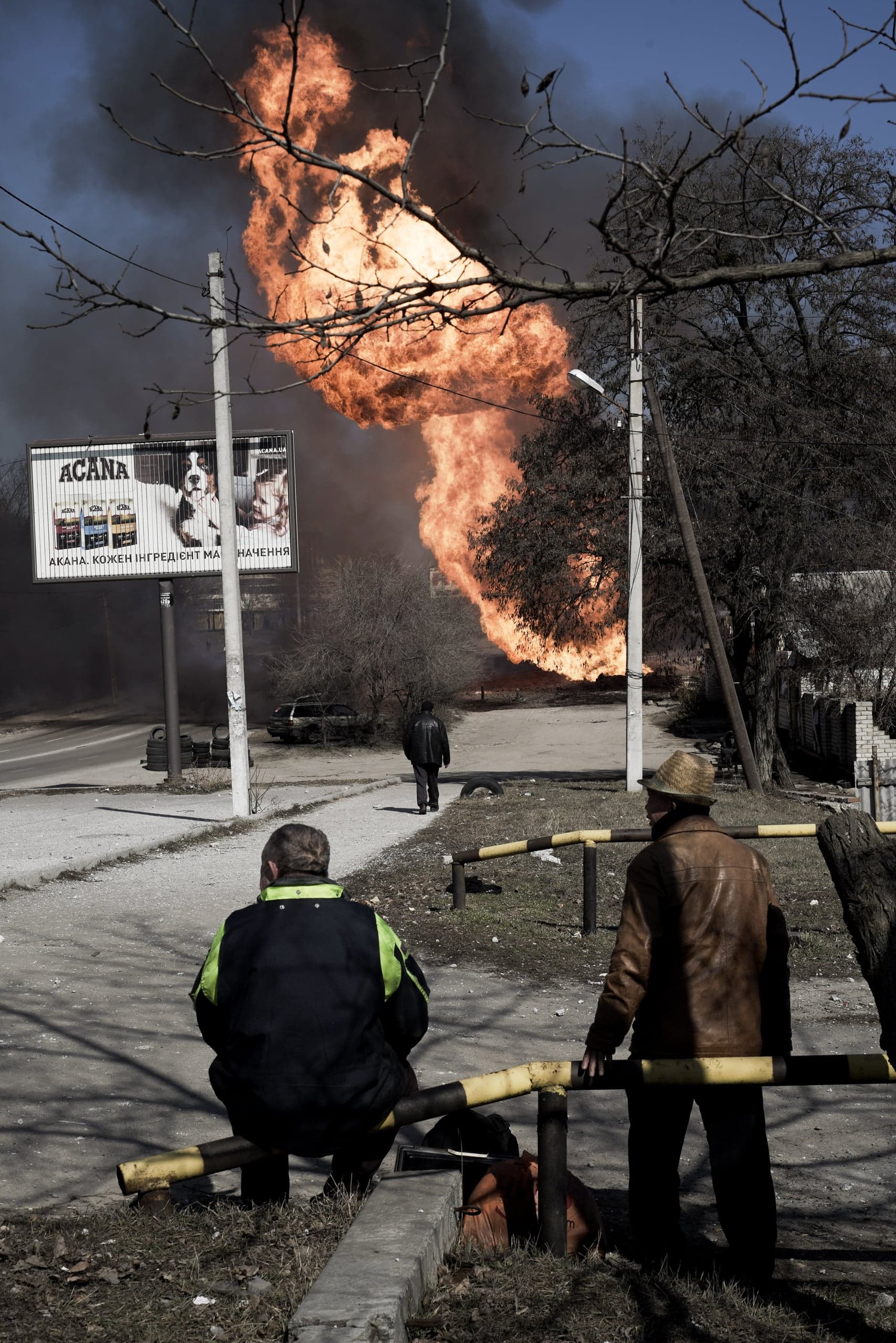 Уличный снимок: двое мужчин на переднем плане наблюдают за огромным столбом оранжевого пламени и черного дыма, поднимающимся в небо за рекламным щитом с рекламой корма для животных.