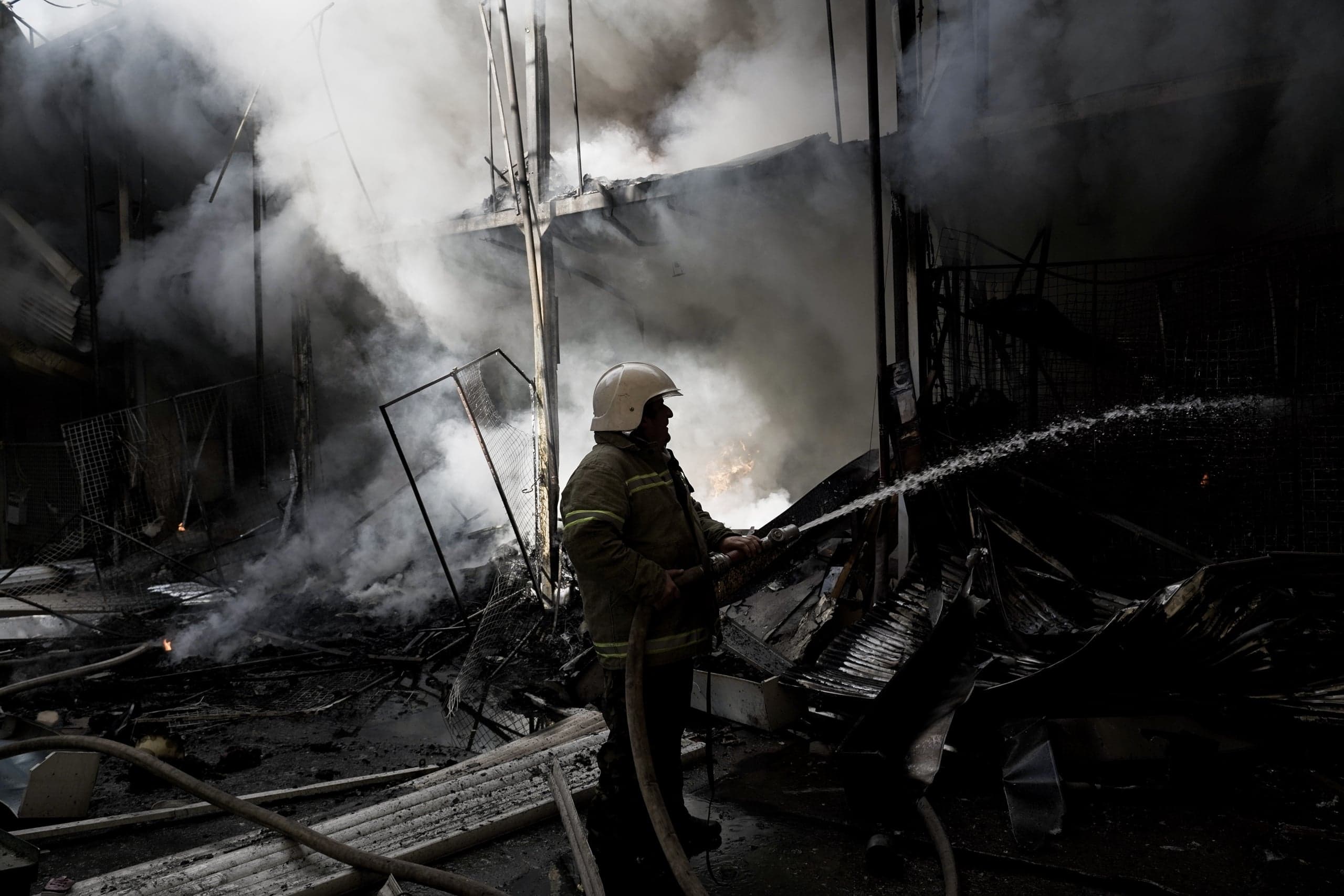 Пожарный в каске тушит водой обгоревшие руины здания, окруженные густым дымом.