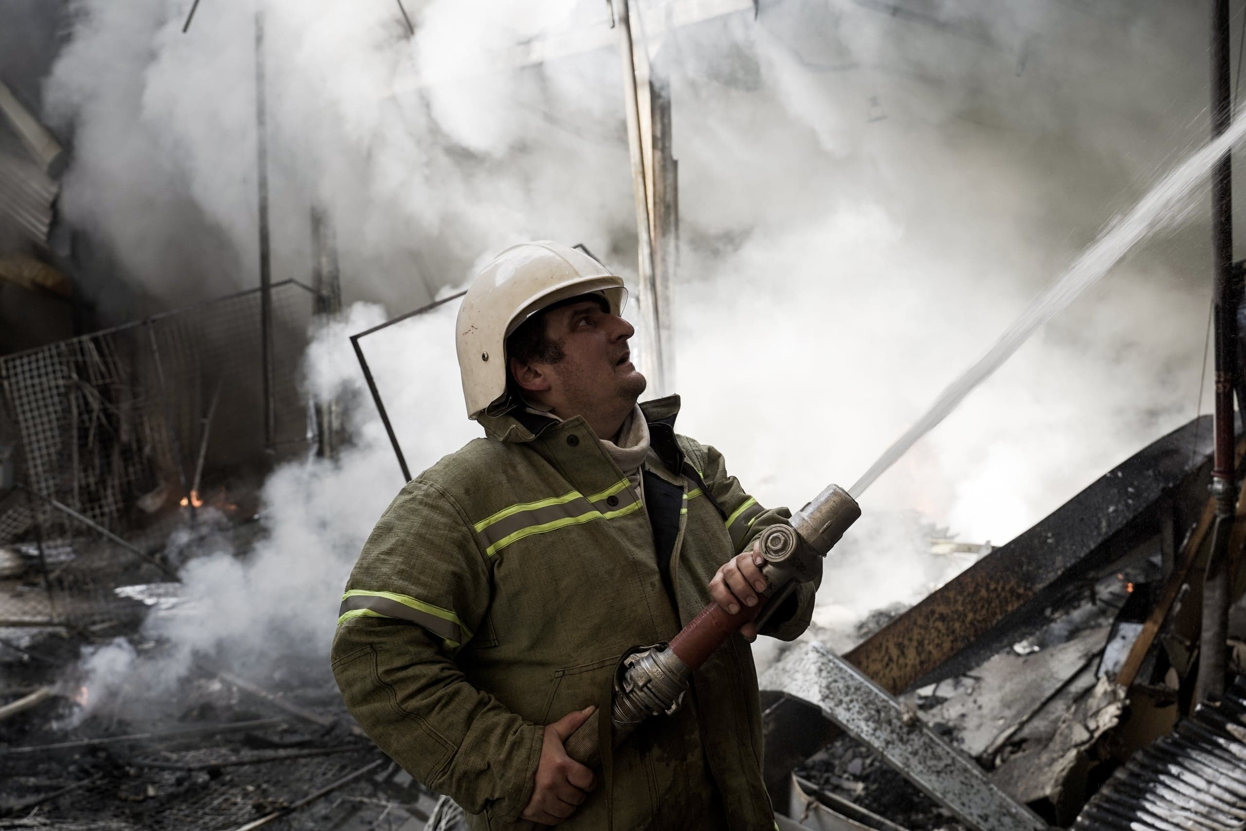 Пожарный в защитном костюме и каске направляет струю воды из брандспойта среди густого белого дыма и обломков.