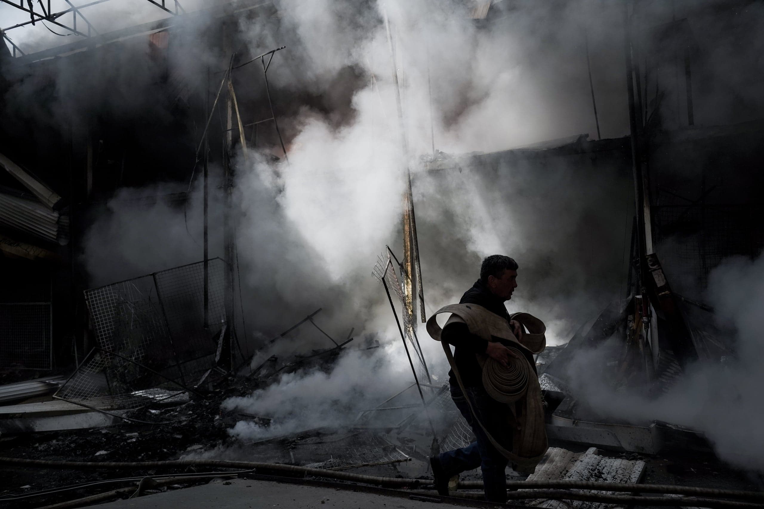 Человек несет свернутый пожарный рукав, проходя через густой дым и обломки внутри сгоревшего здания.