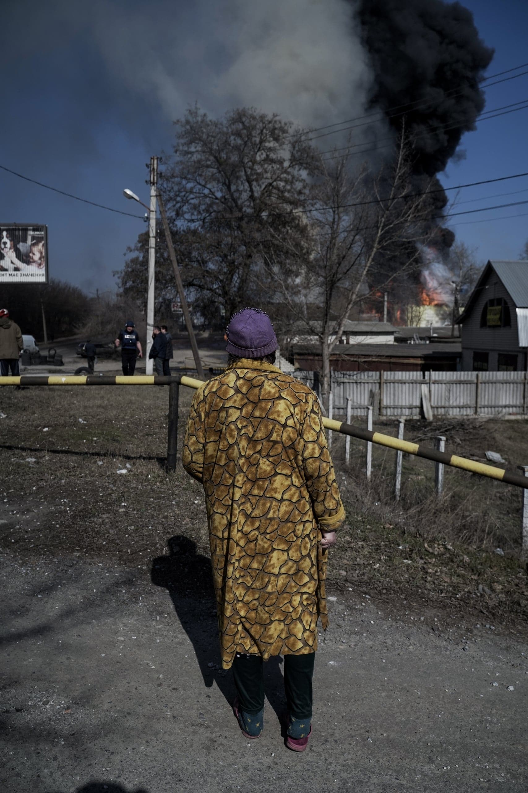 Человек в желто-коричневом пальто и фиолетовой шапке стоит спиной к камере, наблюдая за сильным пожаром и огромным столбом черного дыма, поднимающимся над зданиями вдали.