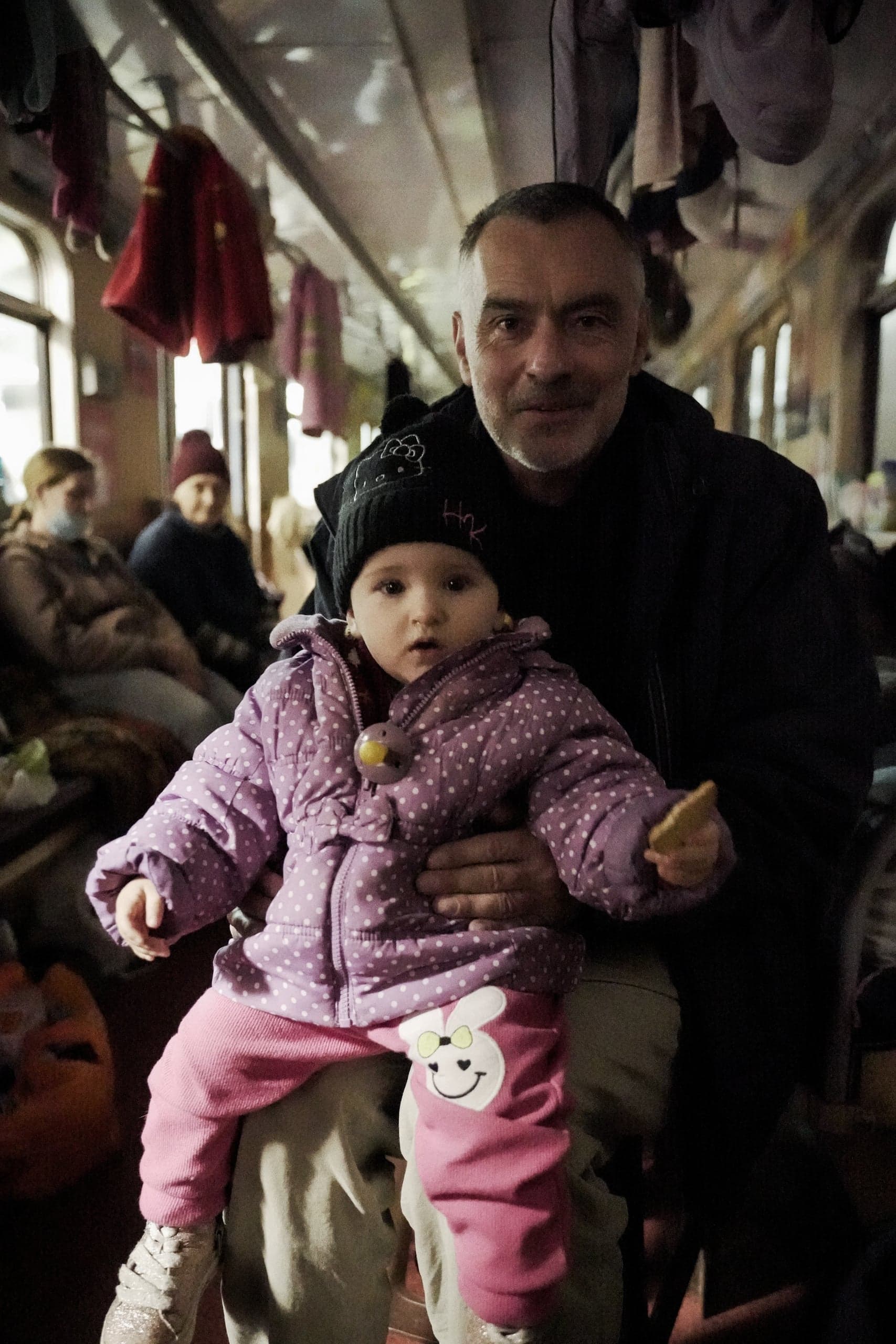 Мужчина держит на руках маленькую девочку в сиреневой куртке в горошек. Они сидят в переполненном вагоне поезда, где под потолком развешаны вещи.