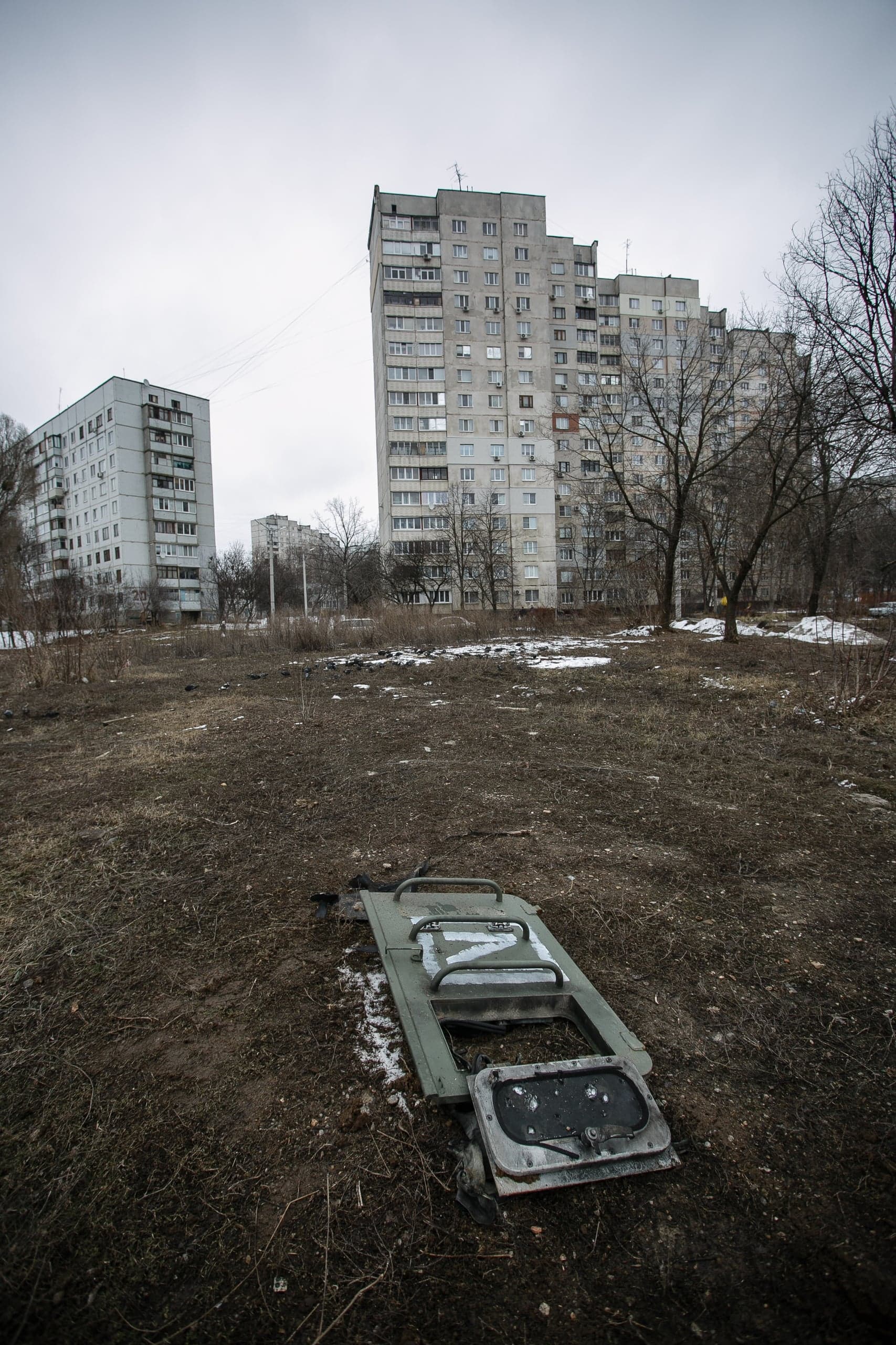 Обломок военной техники с символом «Z» лежит на грязной земле. На заднем плане — высокие серые жилые дома.