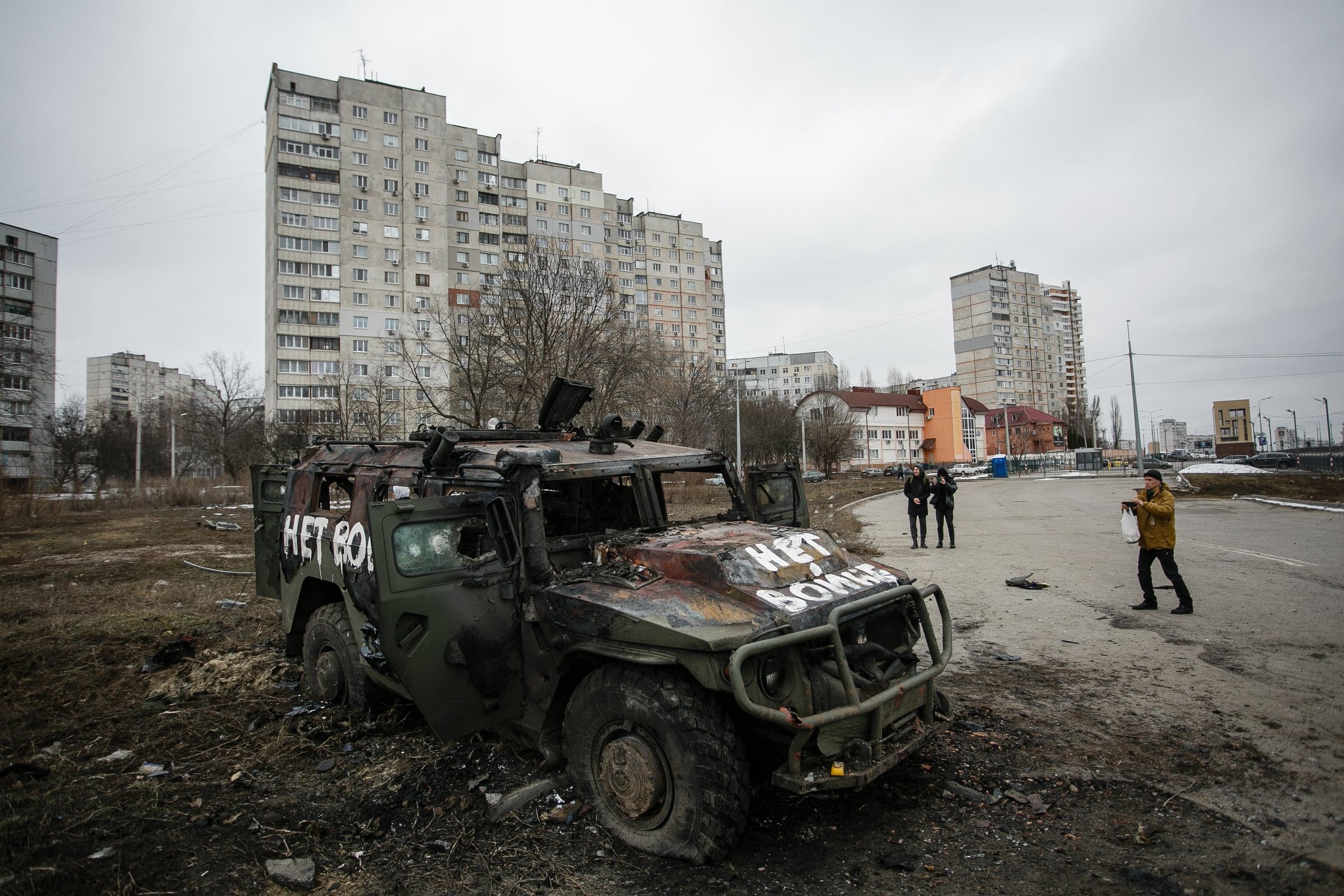 Обгоревший военный броневик с надписью "НЕТ ВОЙНЕ" на фоне жилых многоэтажных домов. Видны прохожие.