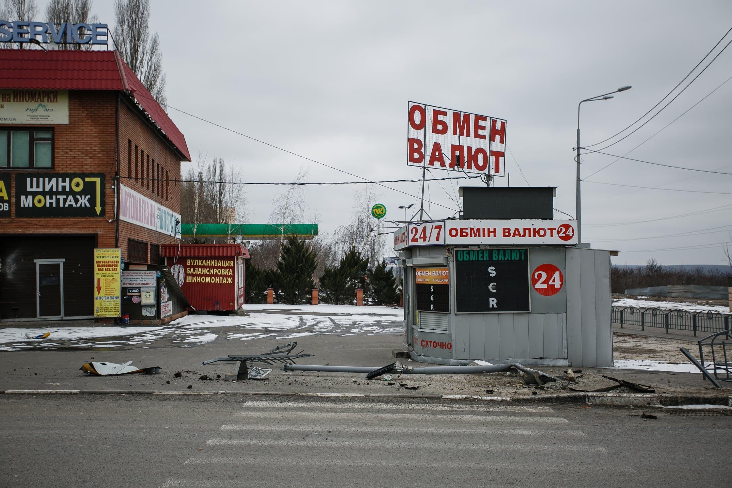 Поврежденный киоск "ОБМЕН ВАЛЮТ" и автосервис. На асфальте лежат обломки, включая части упавшего белого беспилотника.