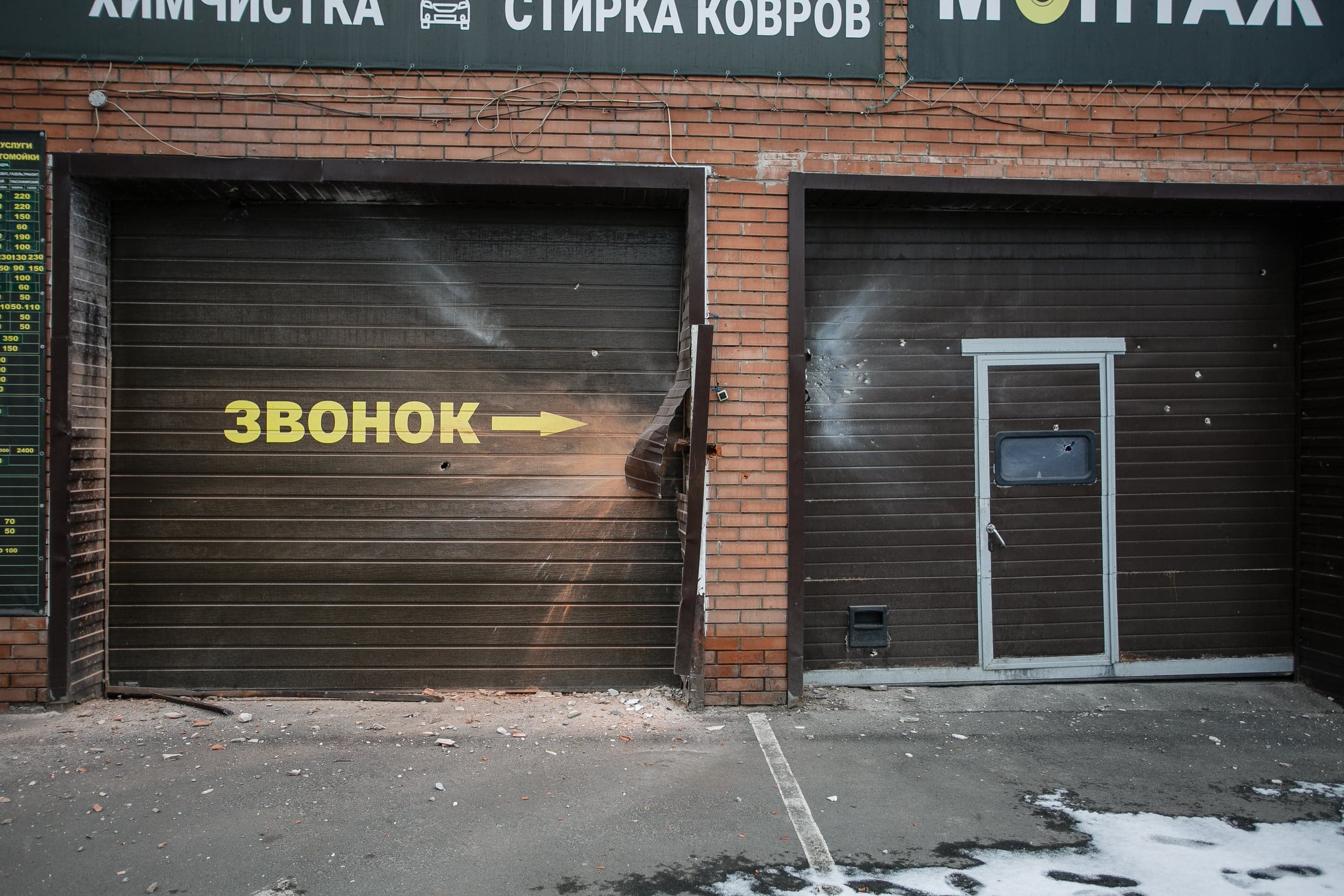 Двое поврежденных обстрелом коричневых гаражных ворот. На левых воротах желтая надпись "ЗВОНОК" со стрелкой, указывающей вправо. Видны пробоины и разрушения.