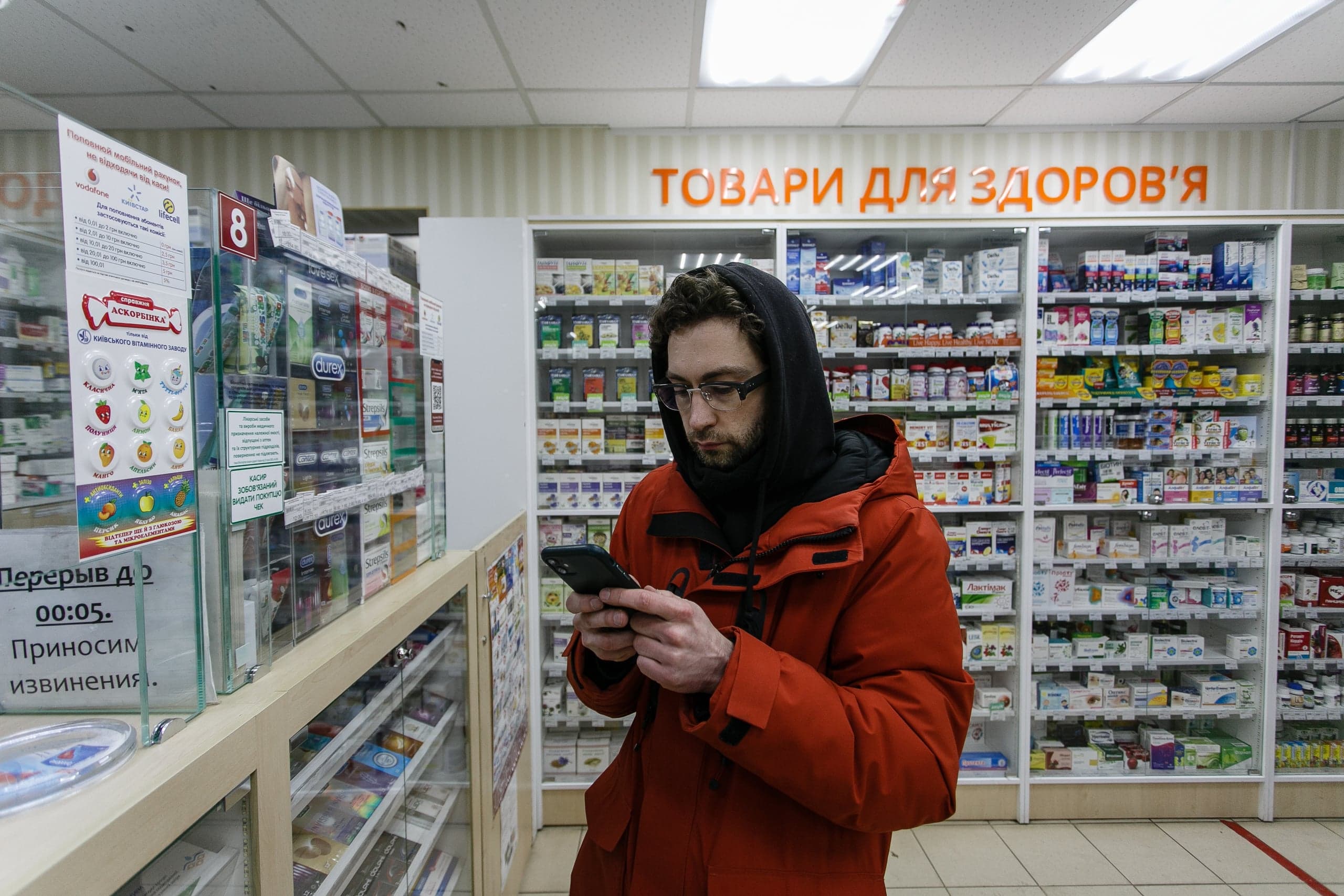 Мужчина в оранжевой куртке и черном капюшоне смотрит в телефон в отделе аптеки на фоне стеллажей с лекарствами. Надпись сверху: "ТОВАРЫ ДЛЯ ЗДОРОВЬЯ".