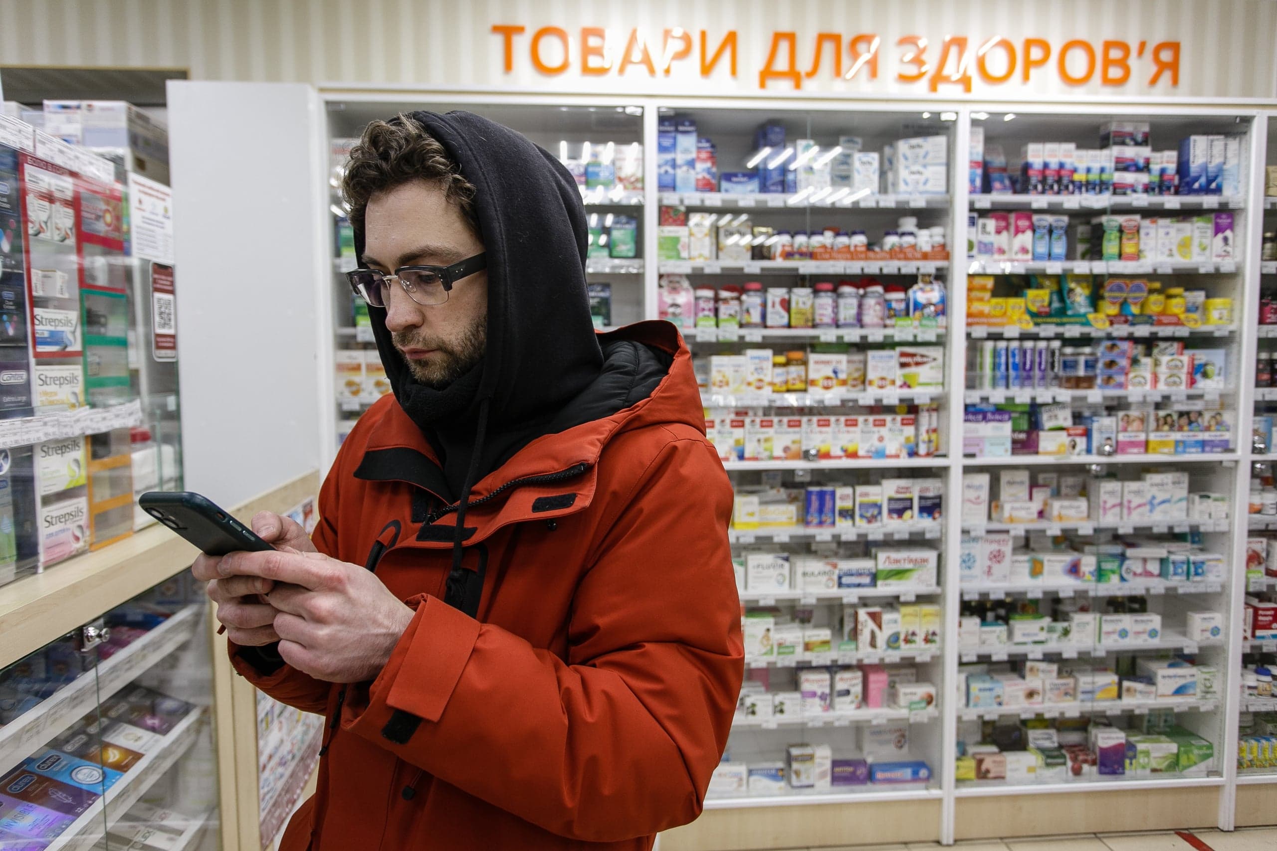 Мужчина в оранжевой куртке и черном капюшоне стоит в аптеке и смотрит в телефон на фоне витрин с лекарствами. Надпись на витрине: "ТОВАРЫ ДЛЯ ЗДОРОВ'Я".